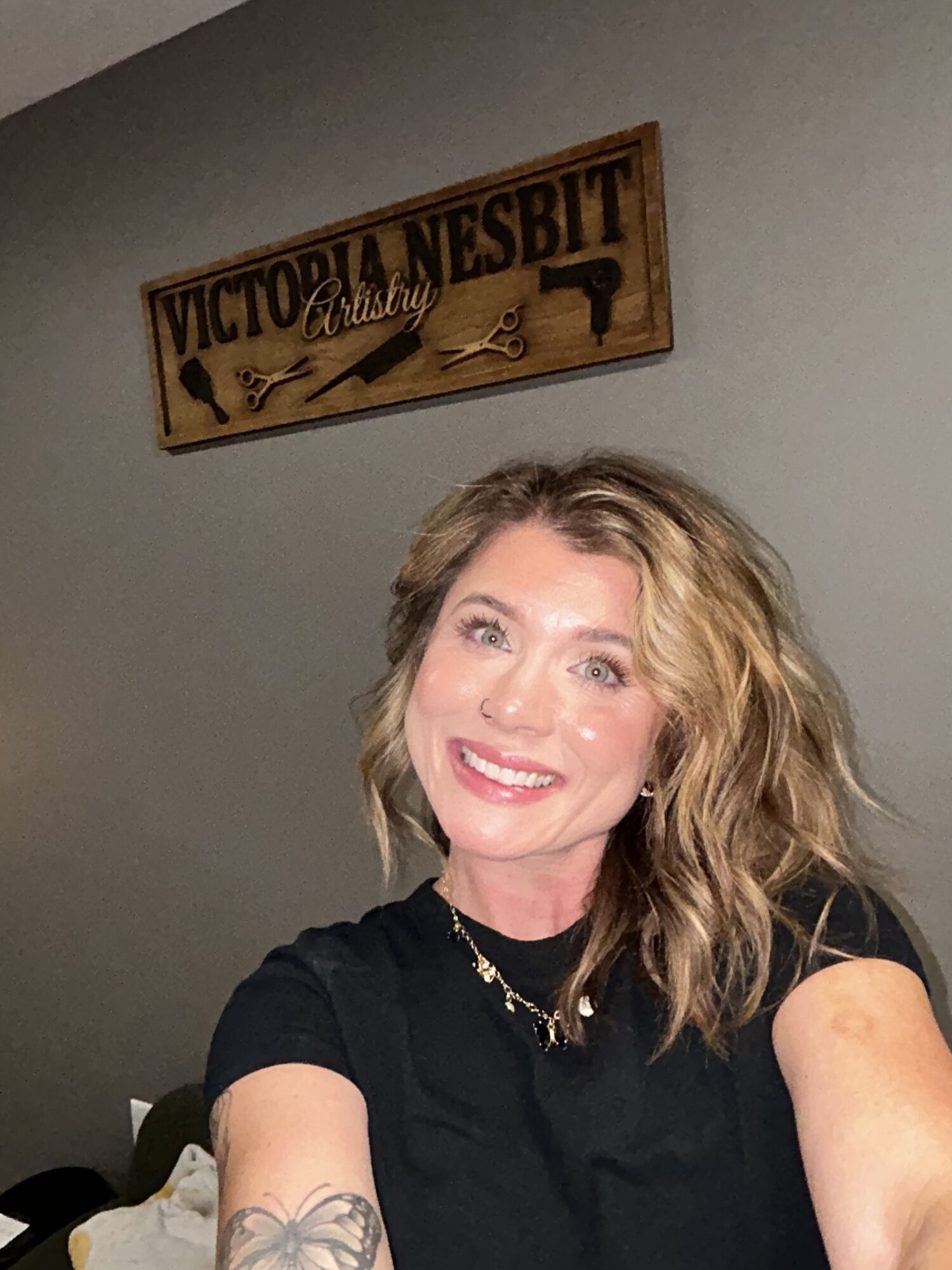Woman with blonde hair smiling, wearing a black shirt and necklace, in front of a wall with a wooden sign.