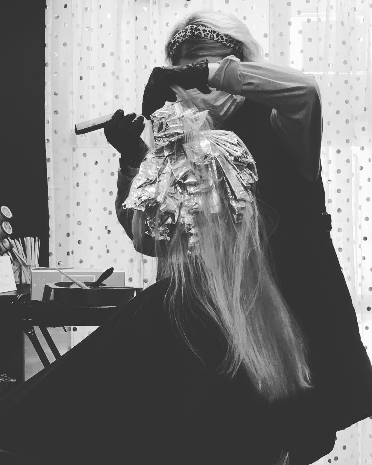 Person applying hair treatment to a seated client in a salon, with hair covered in foil. Black and white image.