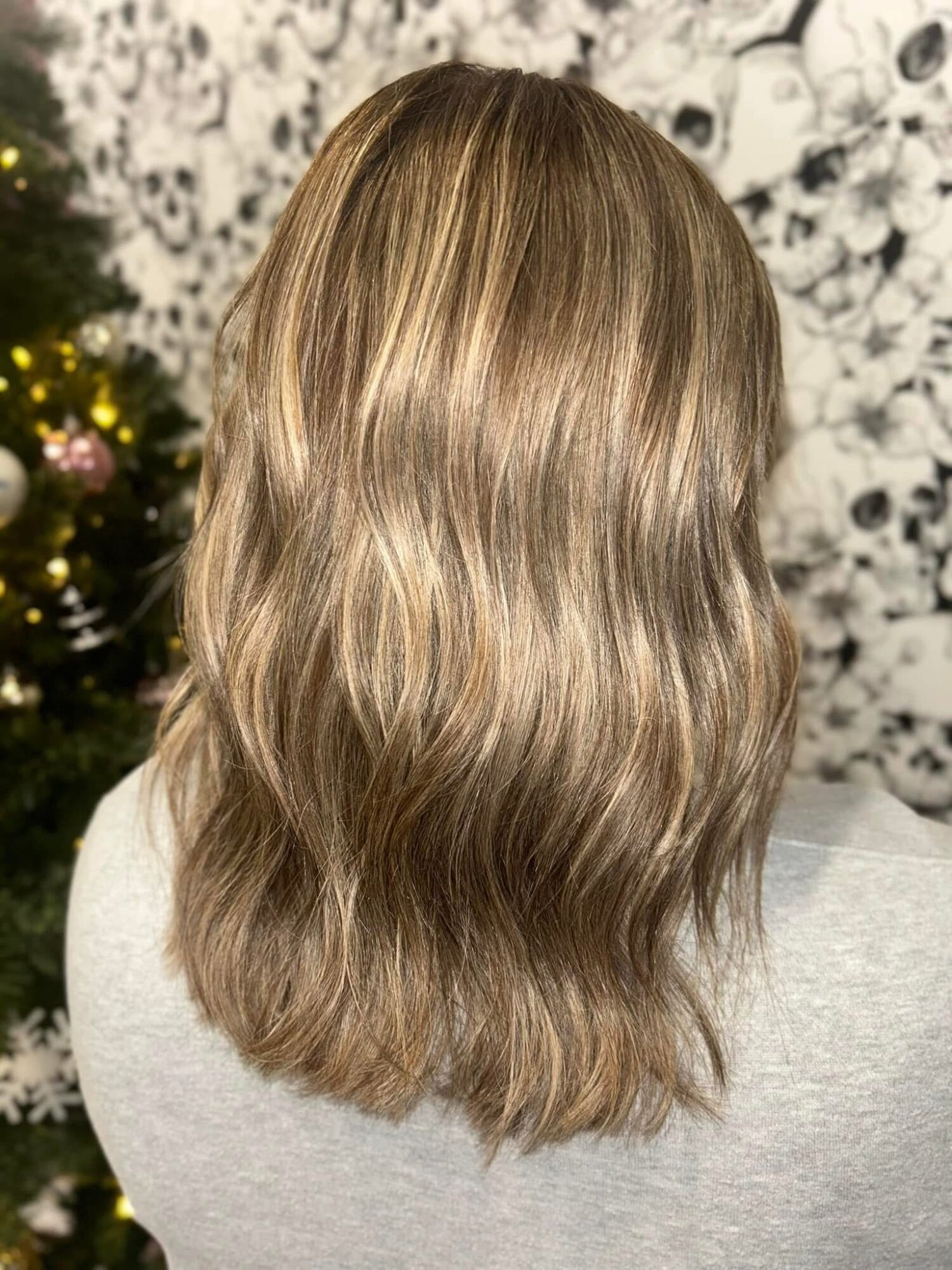 Back of a person with long, wavy, blonde hair, standing in front of a floral background and a decorated Christmas tree.