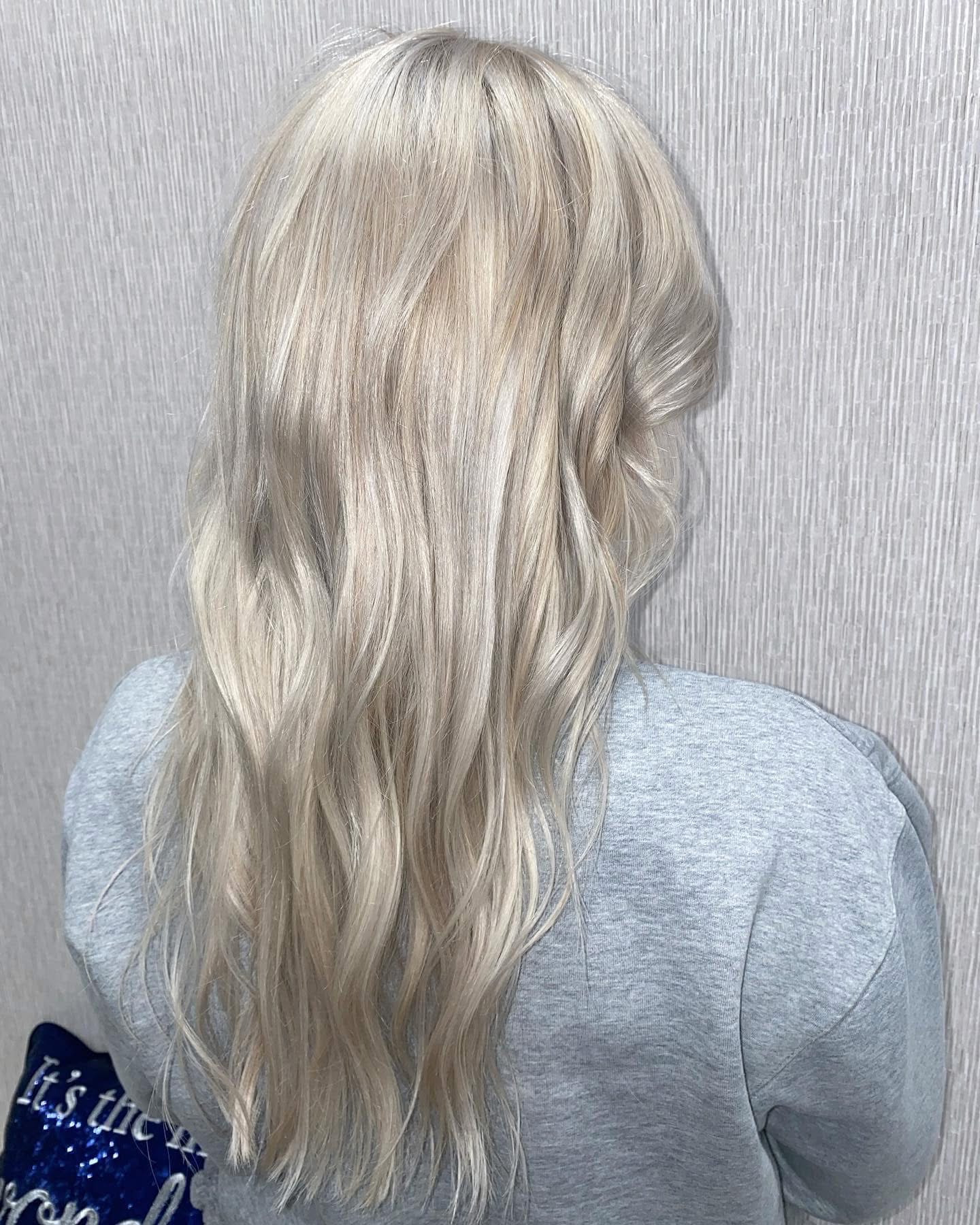 Back view of a person with long, wavy, platinum blonde hair wearing a gray top, standing against a light gray wall.