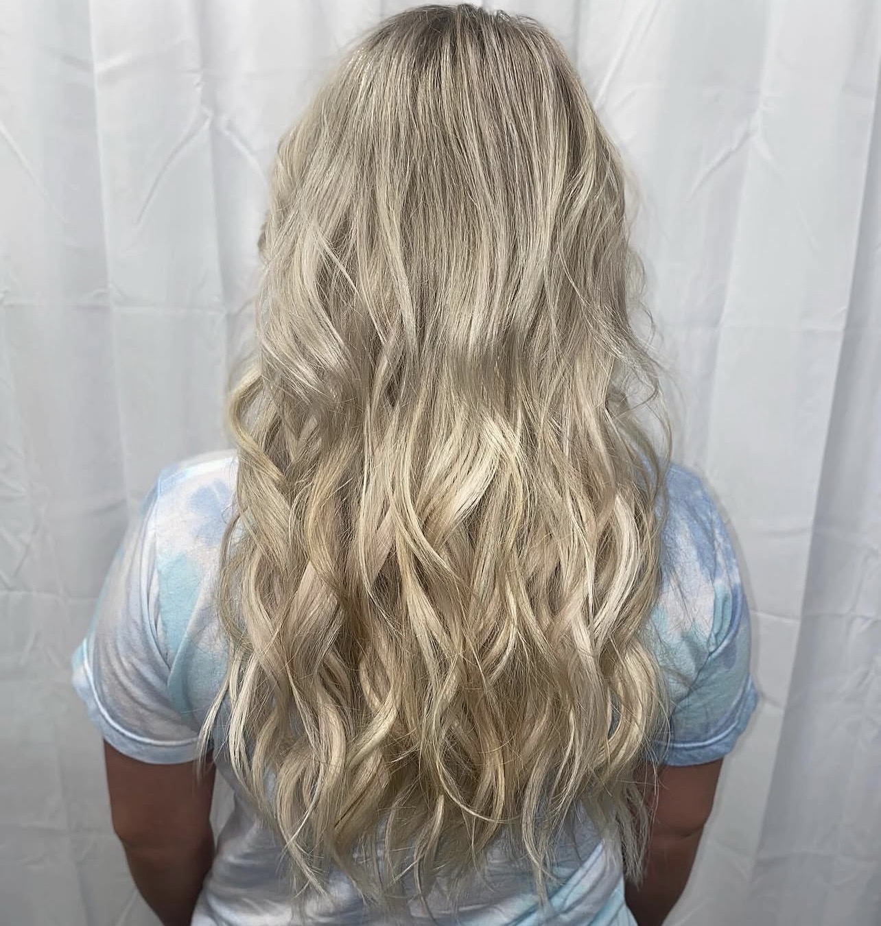 Person with long, wavy blonde hair facing away, wearing a light blue shirt, standing in front of a white curtain.