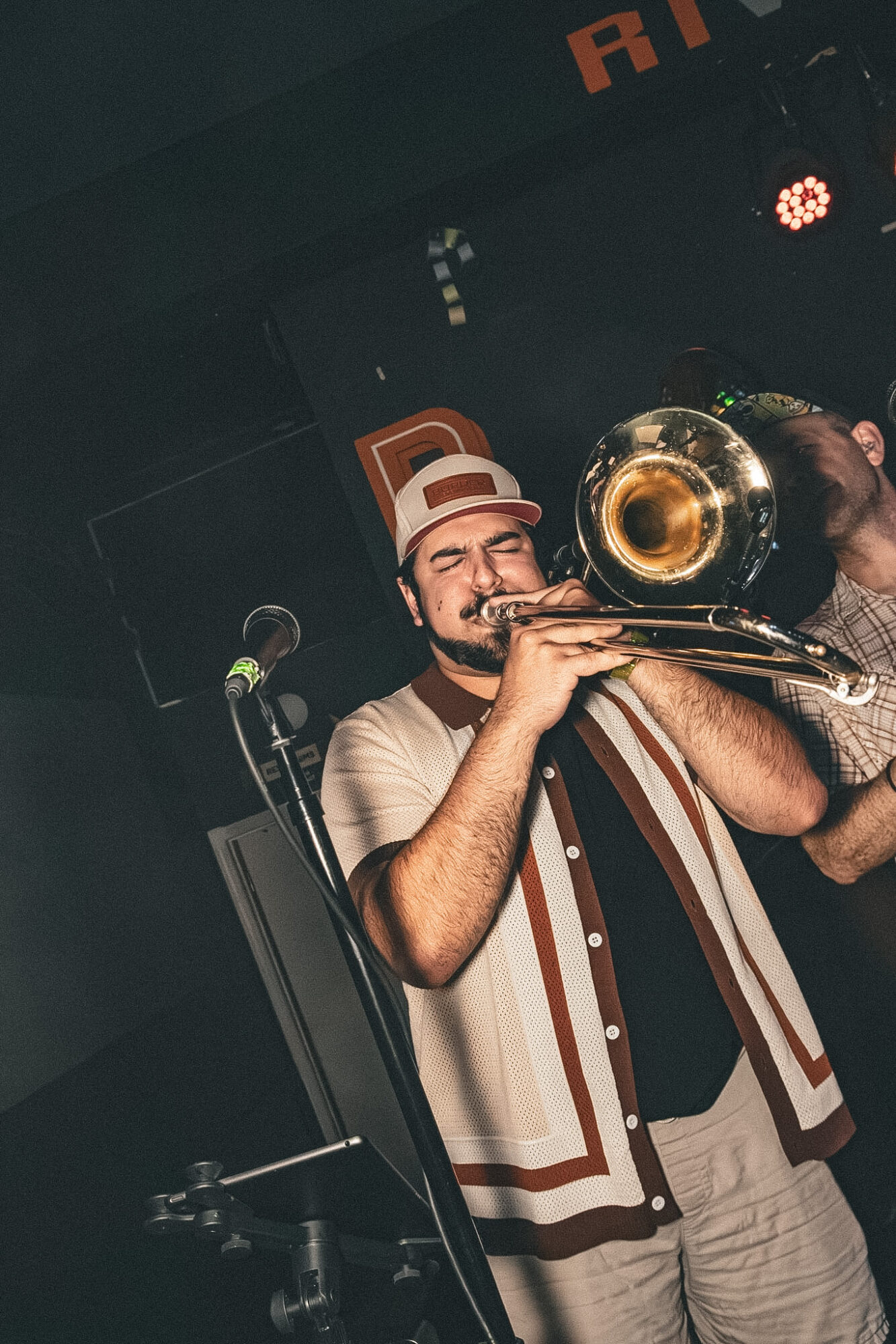 A man playing a trombone on stage, wearing a hat and a patterned vest, with a microphone nearby.