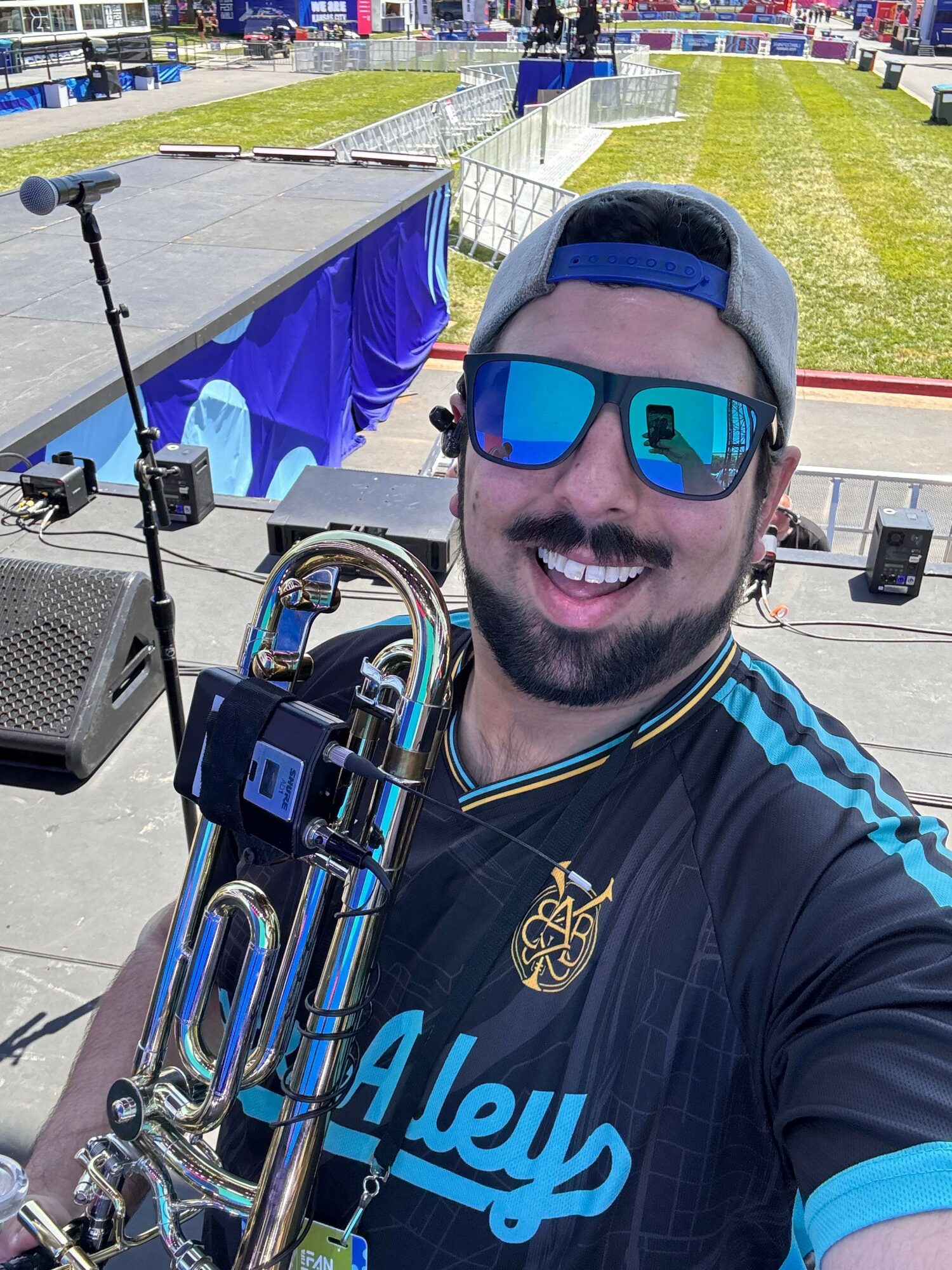 Person smiling with sunglasses holding a trumpet outdoors, wearing a cap and sports jersey.