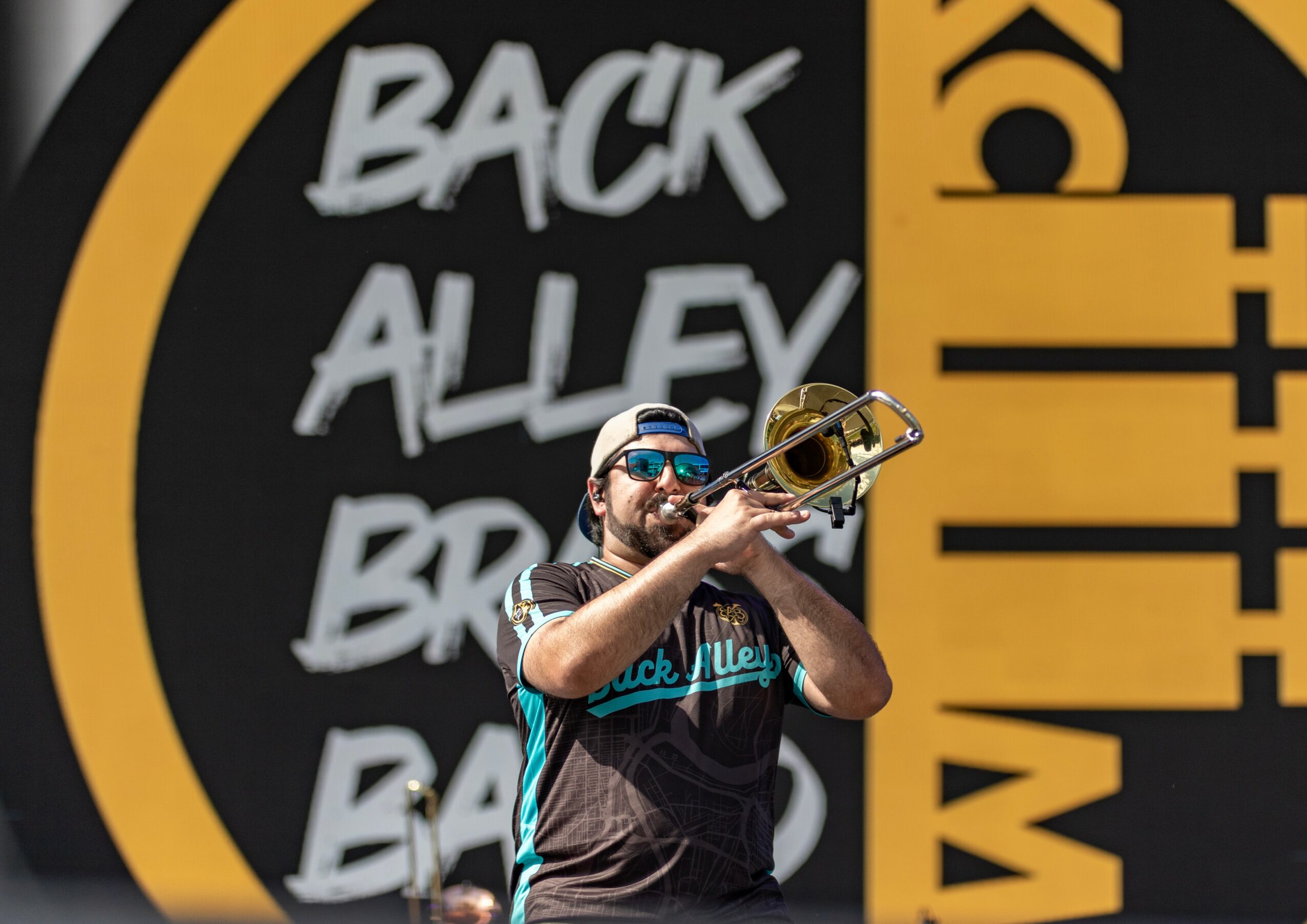 Person playing trumpet outdoors with a large circular sign in the background, reading 'BACK ALLEY' and other text.