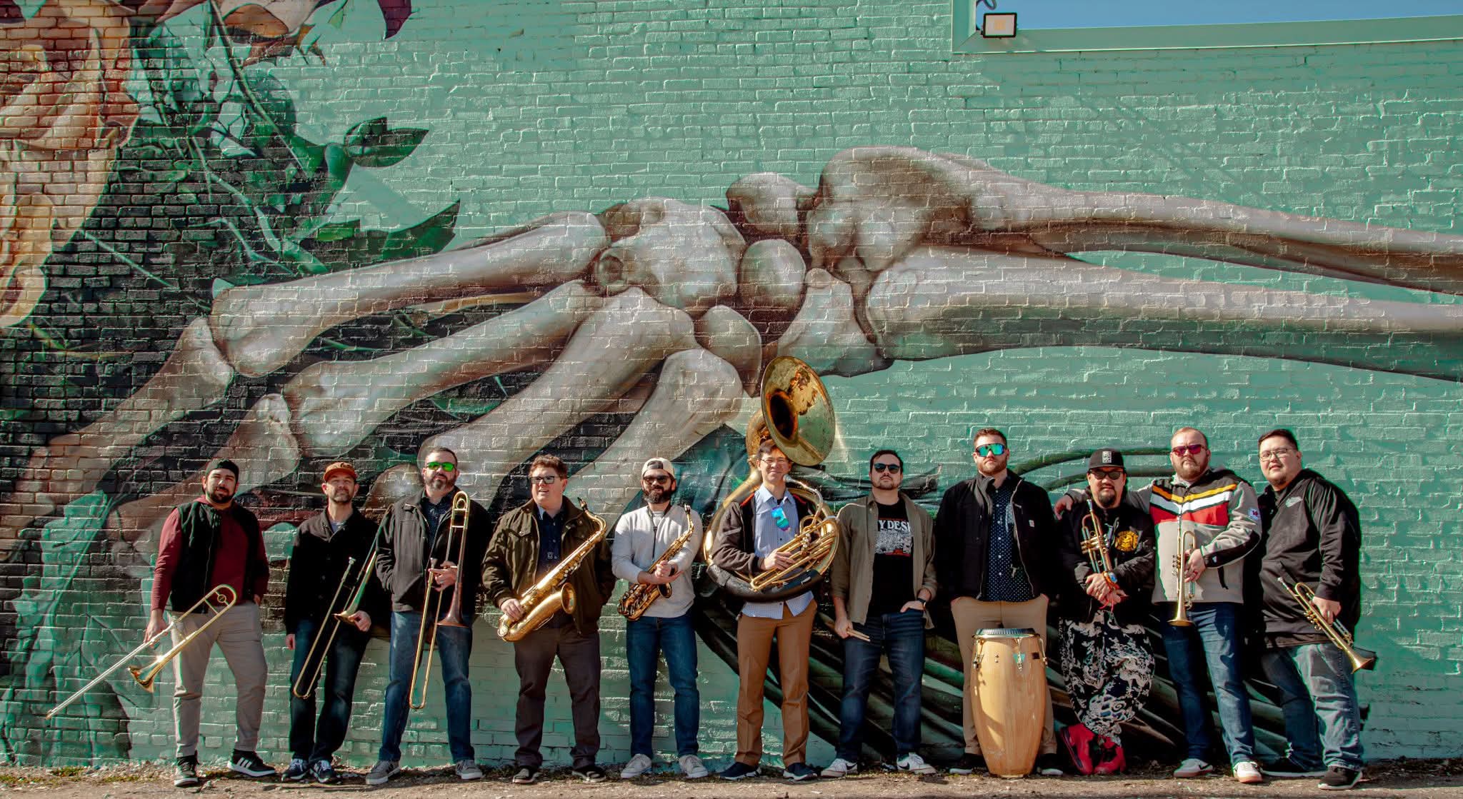 Group of musicians standing in front of a large crab sculpture, holding instruments, with a green background.