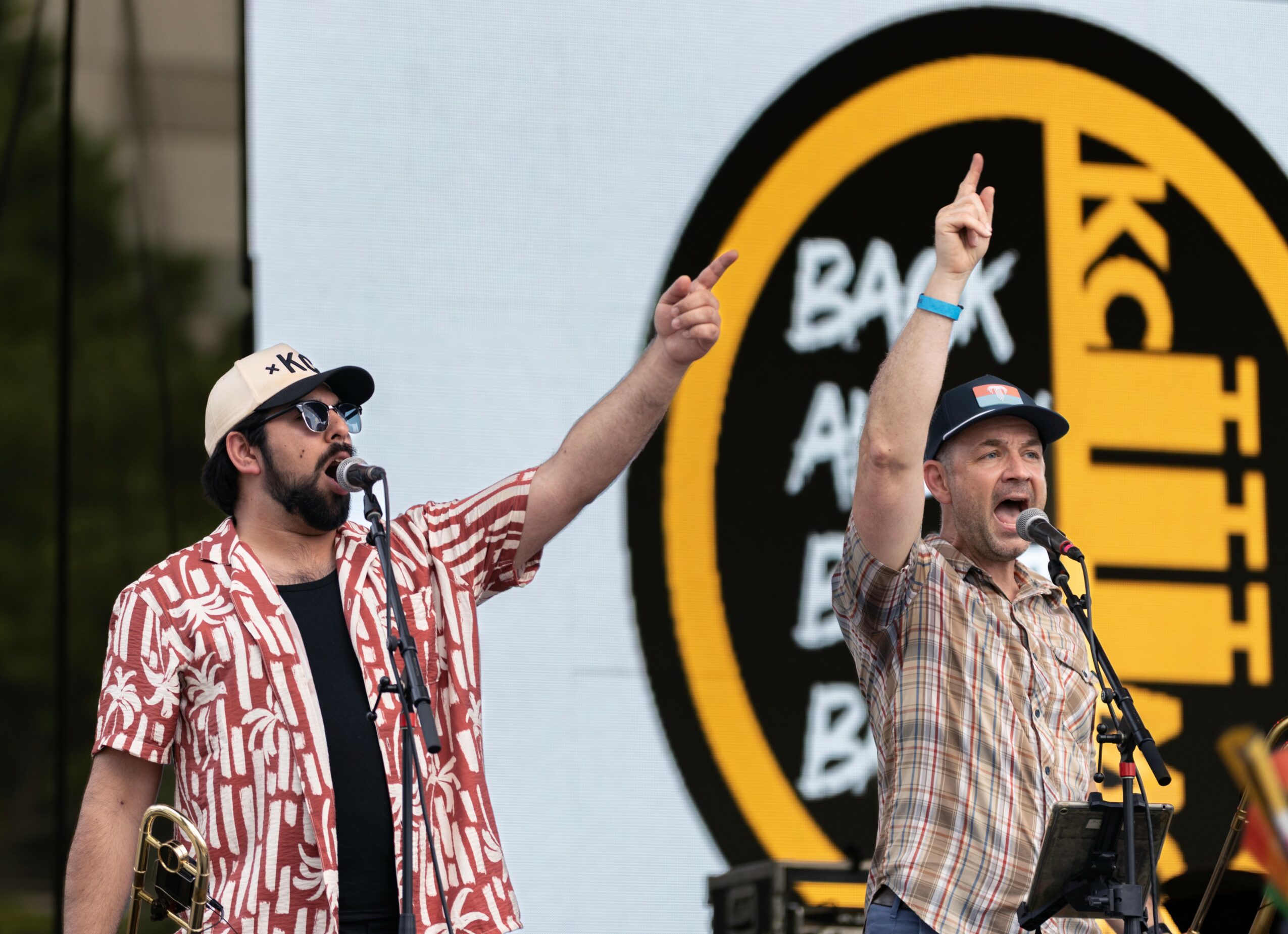 Two men perform on stage with microphones, one wearing a striped shirt and cap, the other in a plaid shirt and cap, in front of a circular sign with text.