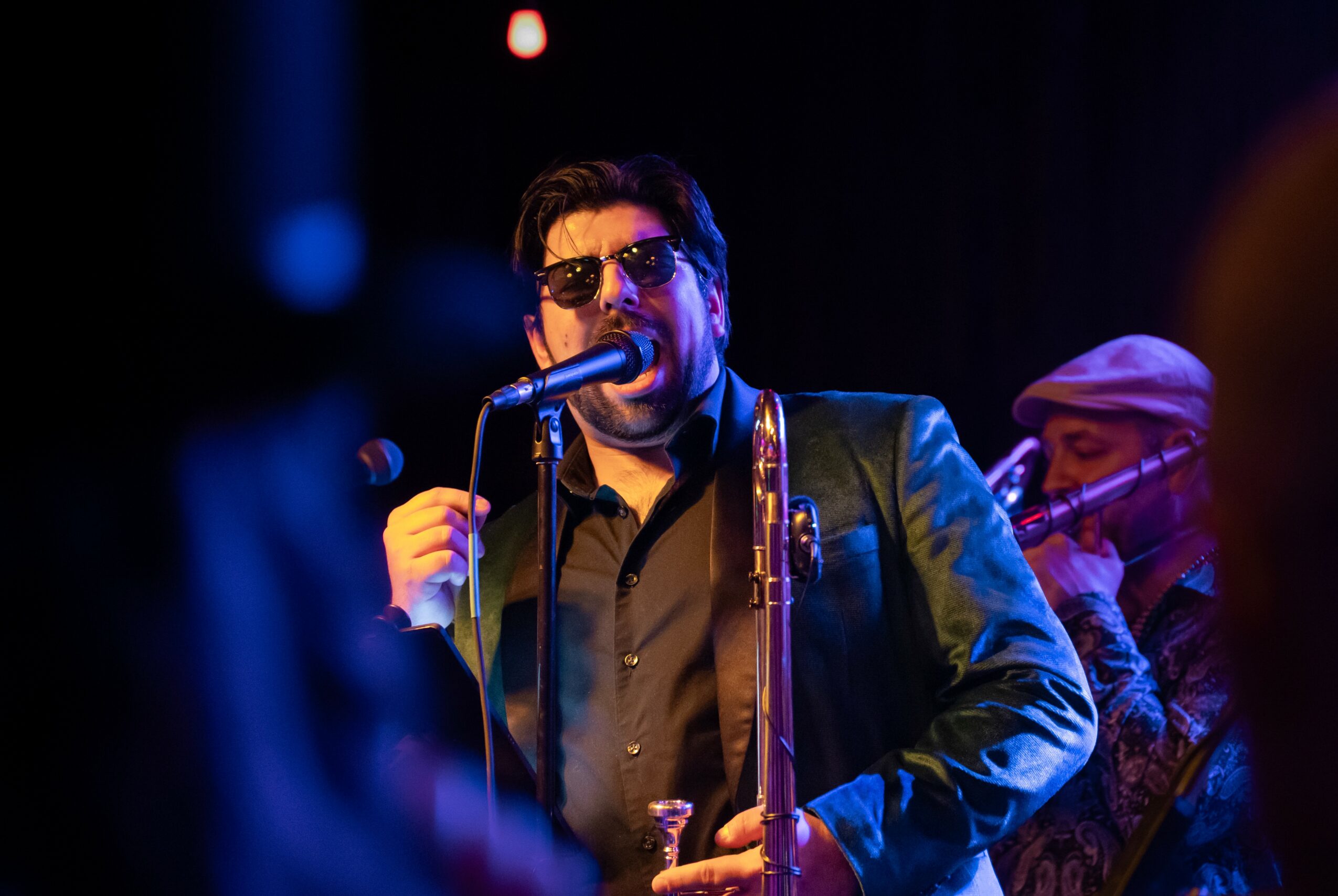 Man singing into a microphone on stage, wearing sunglasses and a leather jacket, with a band member in the background.