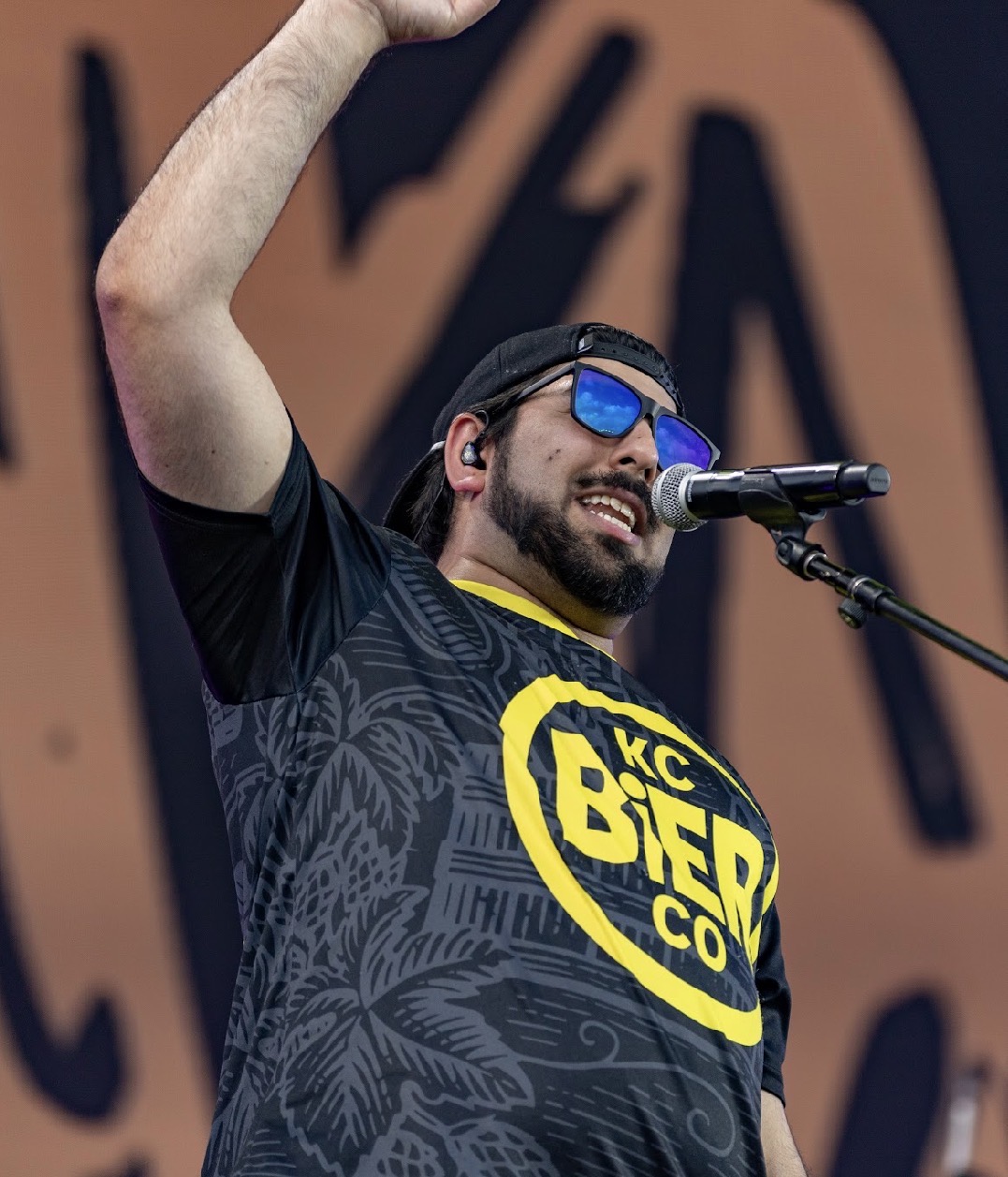 Person with beard and sunglasses speaking into a microphone, wearing a black T-shirt with yellow and black logo, raising one arm.