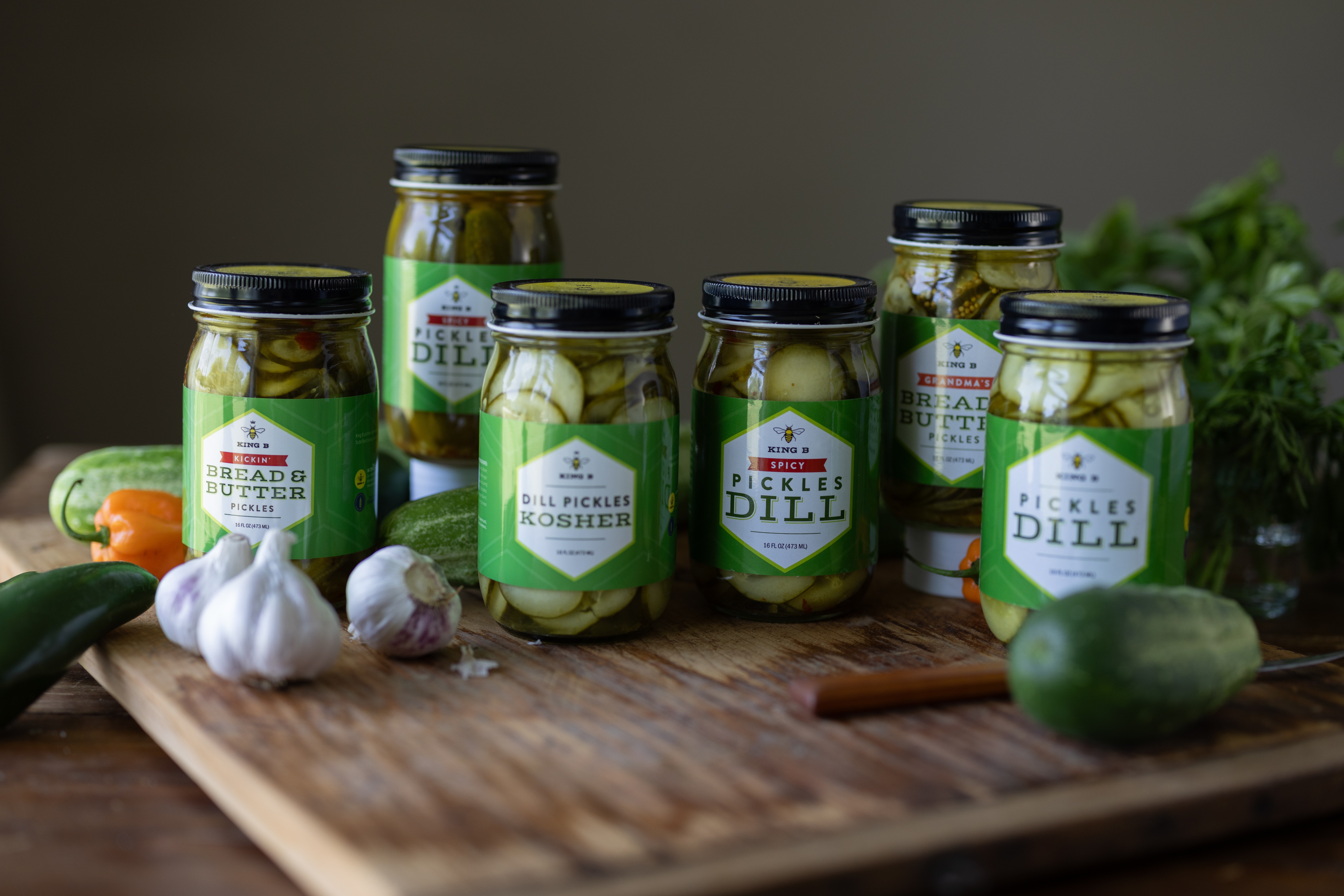 Seven jars of pickles on a wooden cutting board with garlic, cucumber, and herbs, against a dark background.