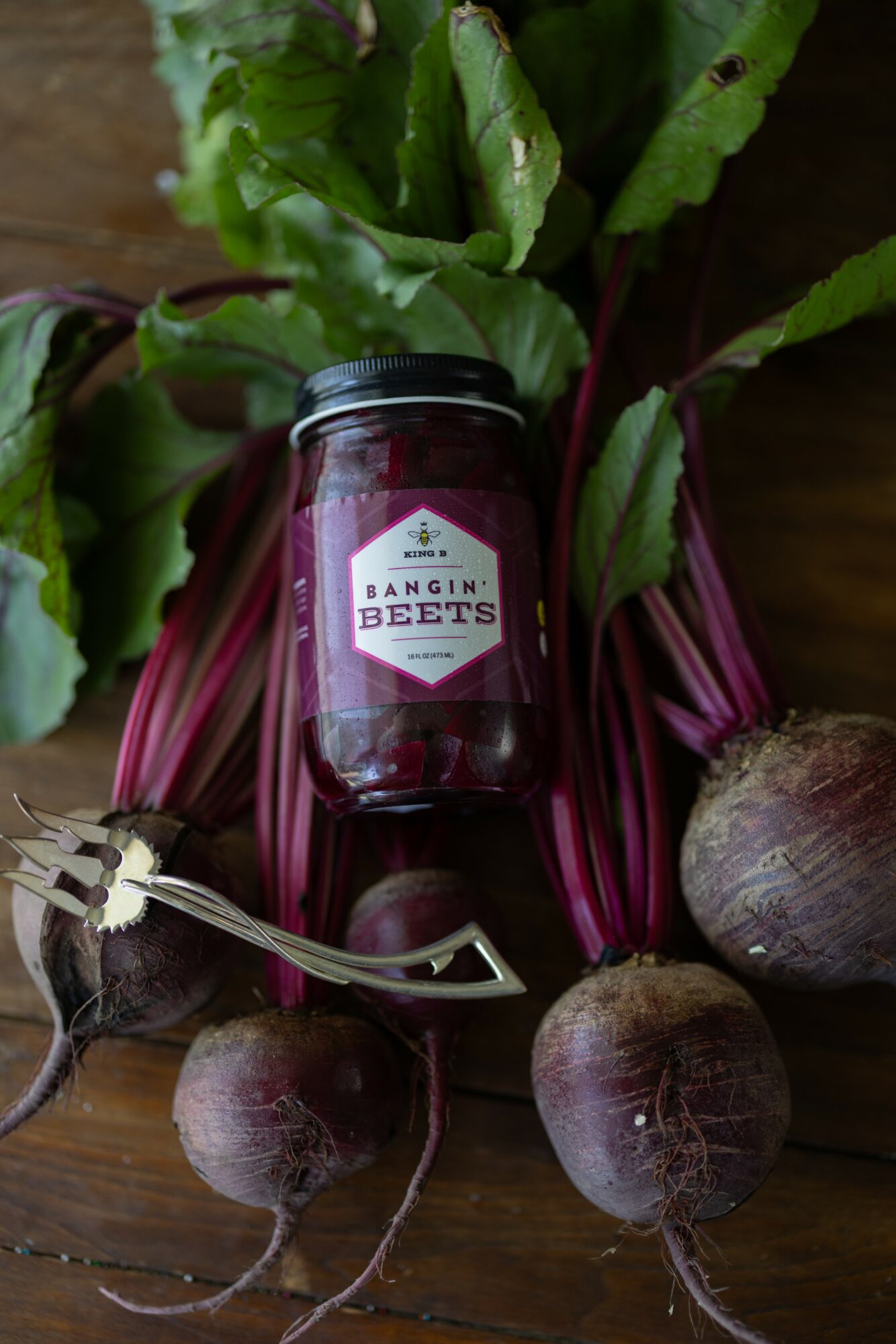 Jar of Bangin' Beets surrounded by fresh beets and green leaves on a wooden surface.