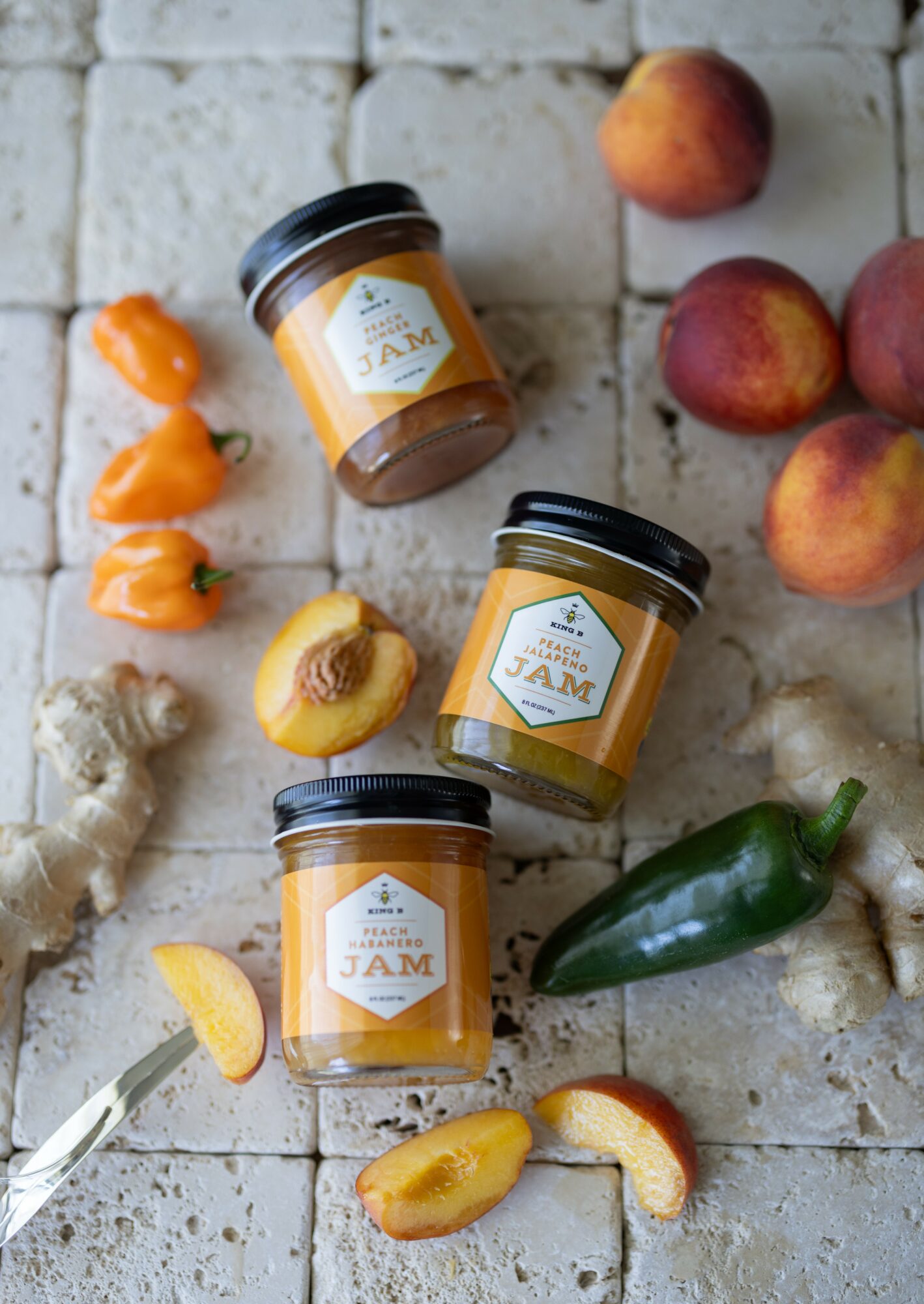 Three jars of jam, peaches, a jalapeño, ginger, and peach slices on a tiled surface.
