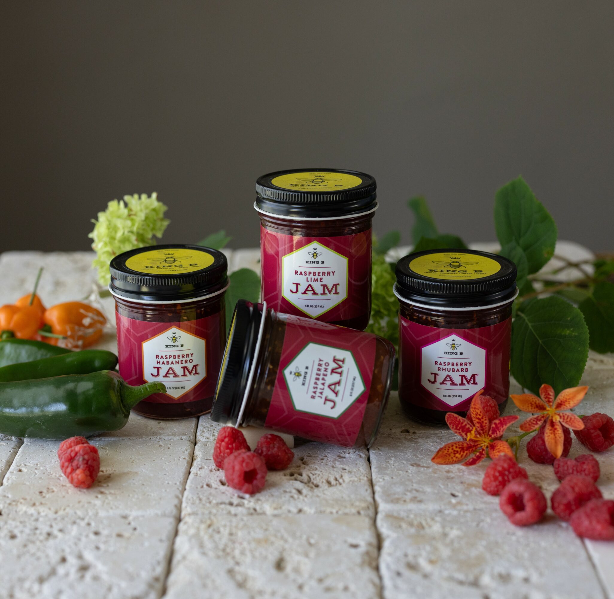 Three jars of raspberry jam surrounded by raspberries, green leaves, and orange peppers on a white wooden surface.