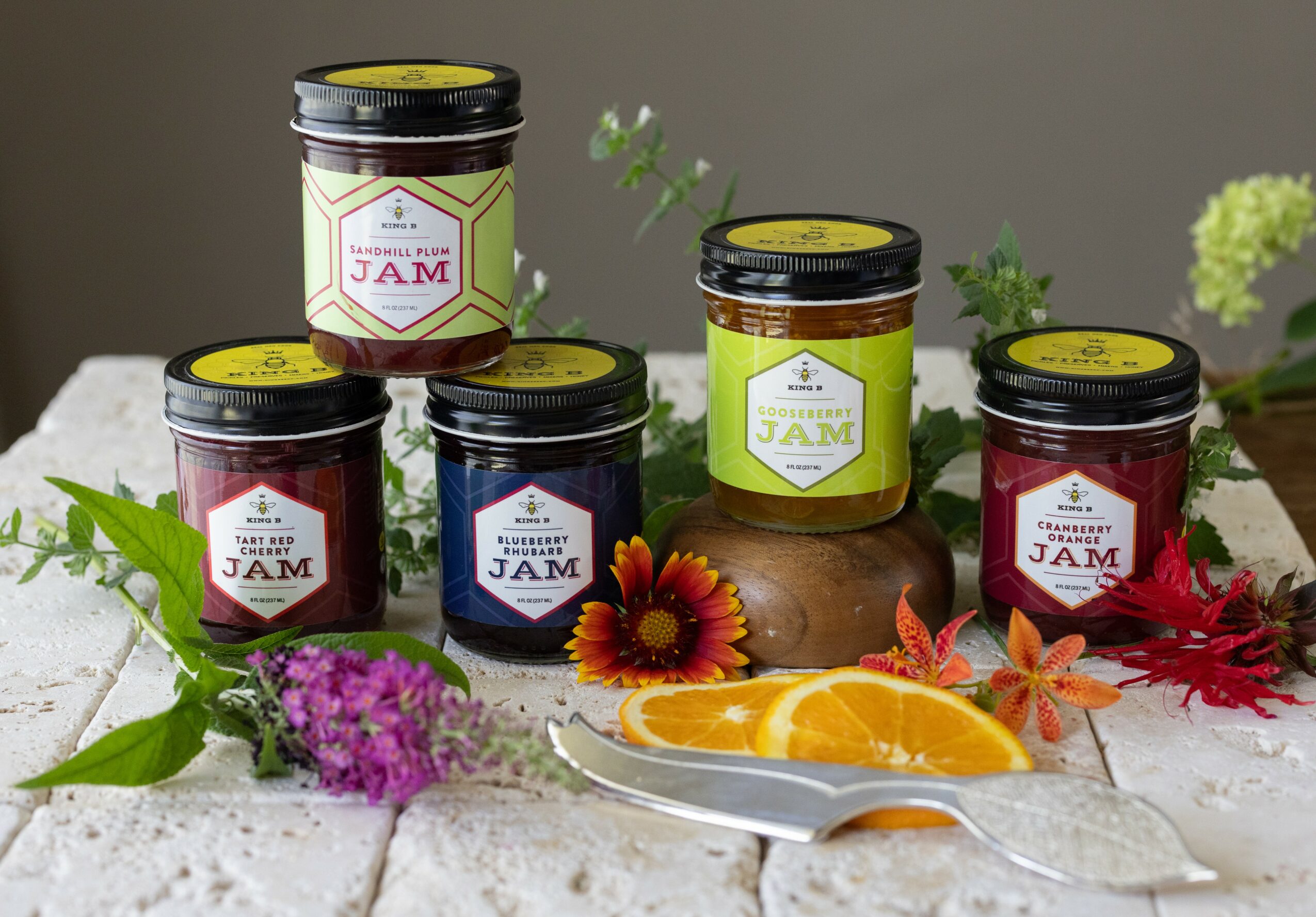 Six jars of jam arranged on a white surface with flowers and orange slices, gray background, some jars have yellow labels.