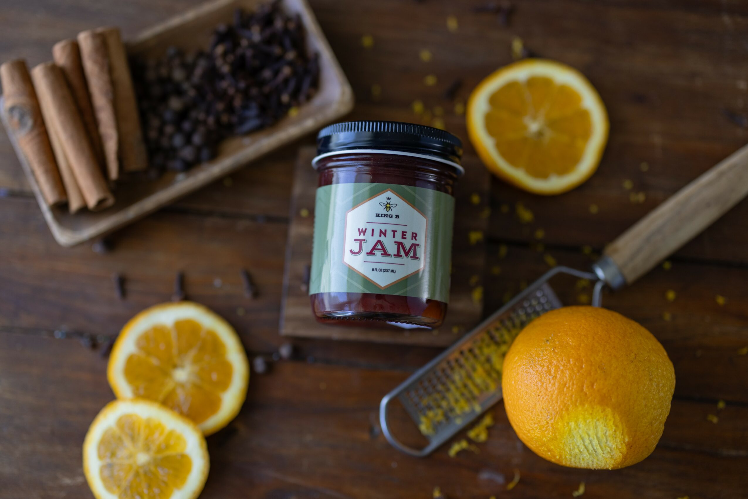 Jar of orange marmalade, sliced oranges, lemon, cinnamon sticks, grater, and a grater on a wooden surface.