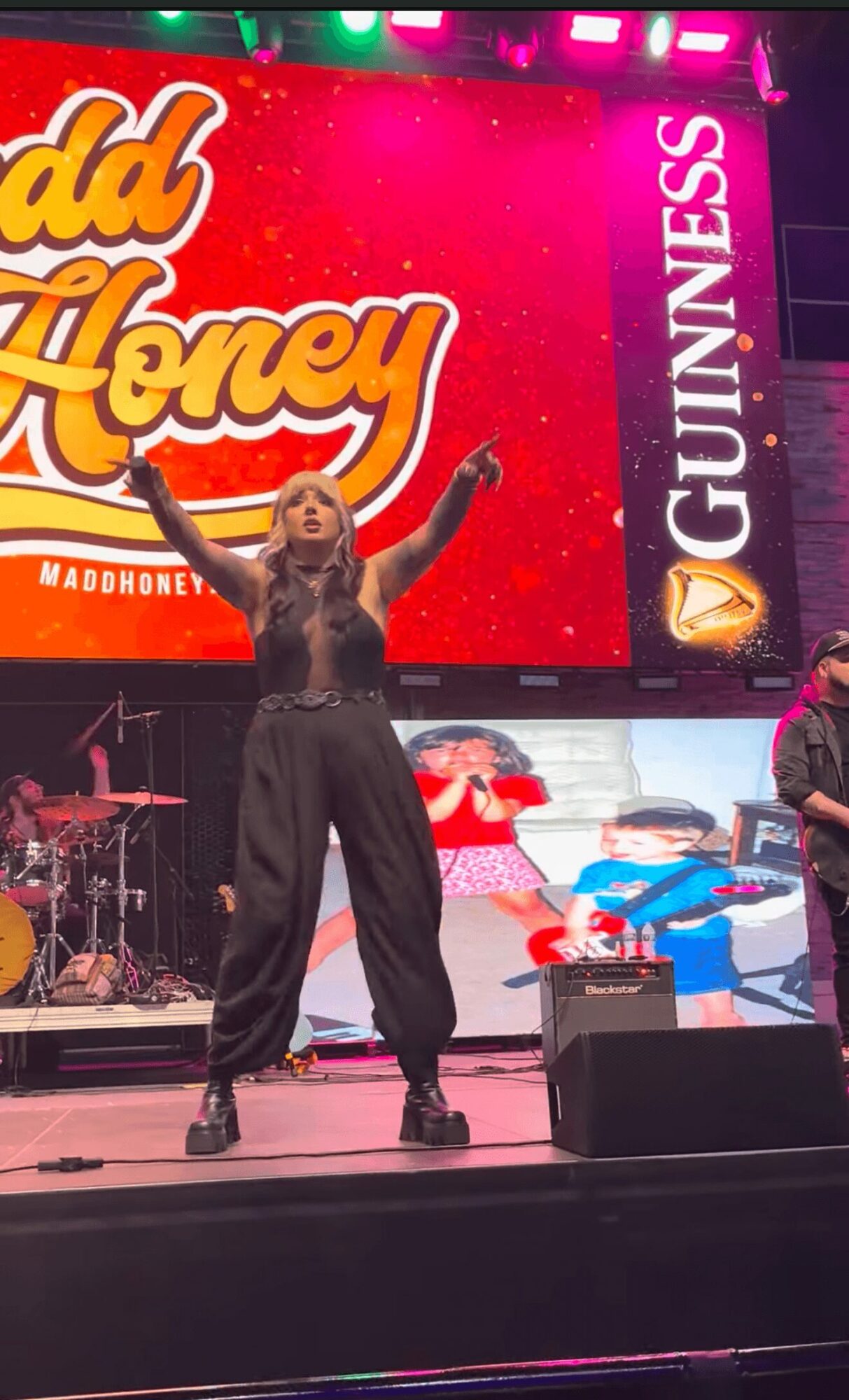 Woman on stage with arms raised, large colorful screen behind her displaying text and logos, musical instruments visible.