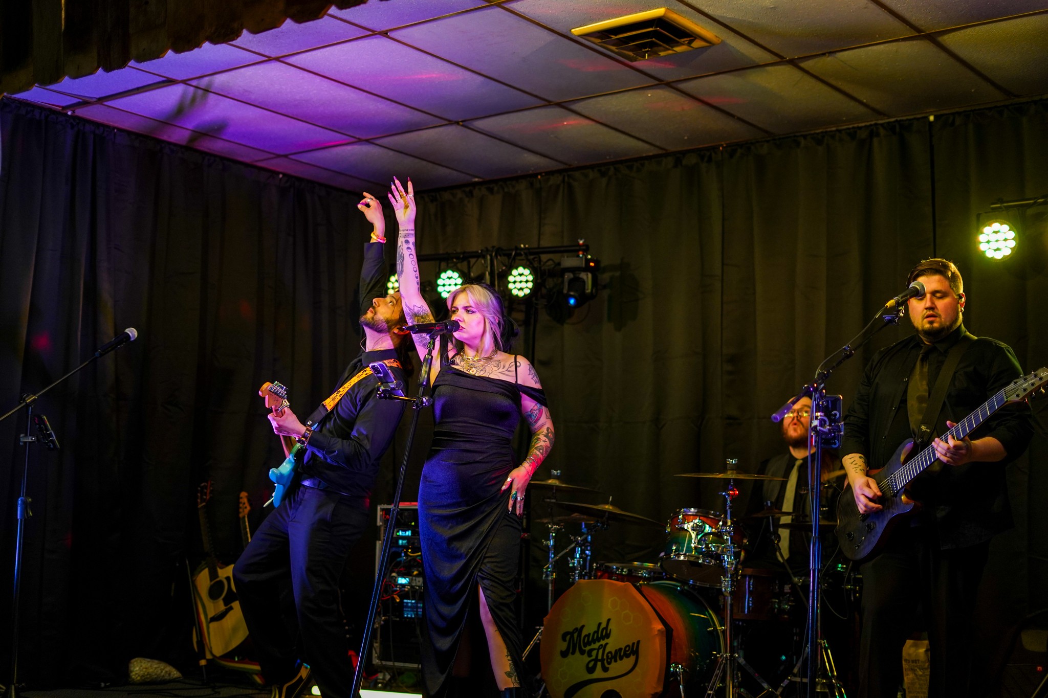 Three performers on stage, one singing into a microphone, one playing guitar, and one raising an arm, with stage lights and instruments in background.