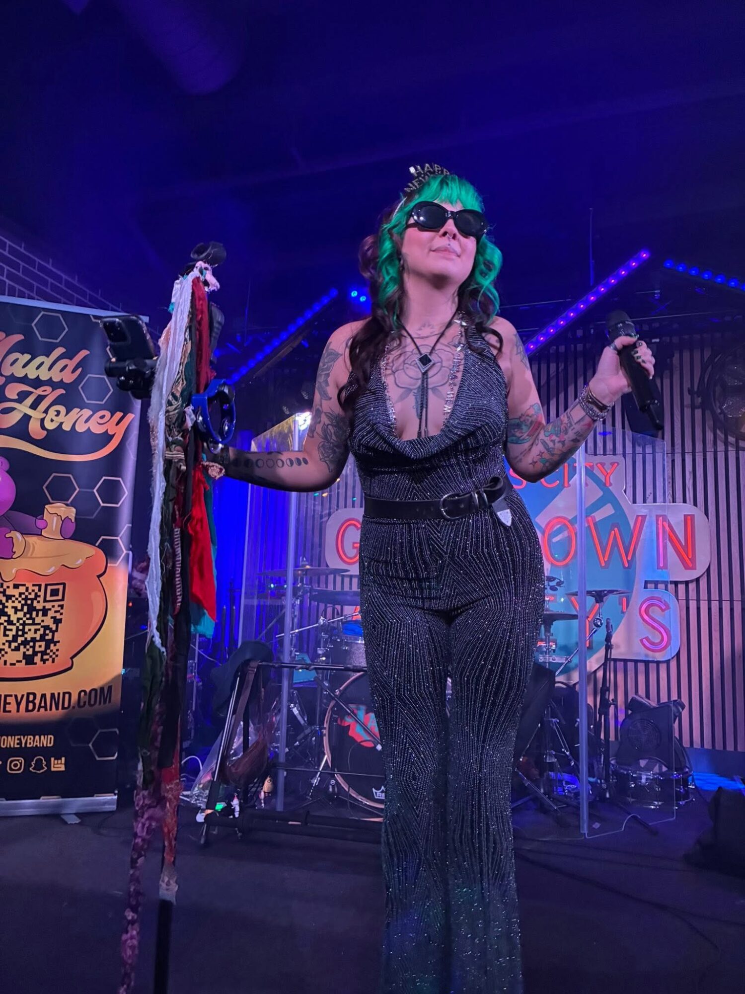 Woman with green hair and sunglasses performing on stage with a microphone, wearing a black outfit, in a concert setting.