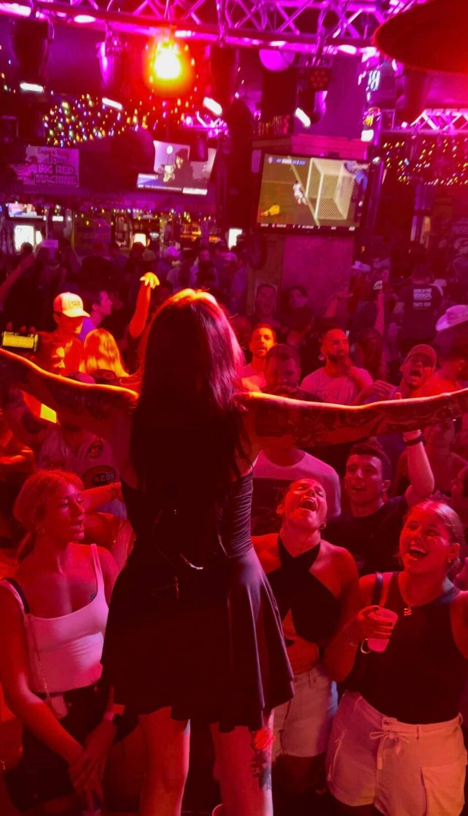 Person with long hair in a black dress stands in a crowd at a lively event with colorful lights.