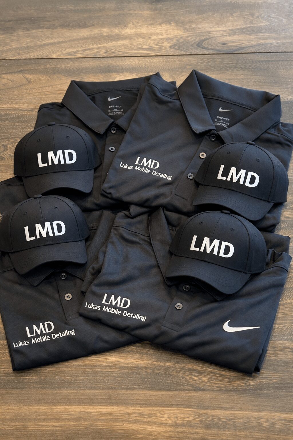 Black polo shirt with LMD logo, six black caps with LMD logo, and a black Nike bag on a wooden surface.