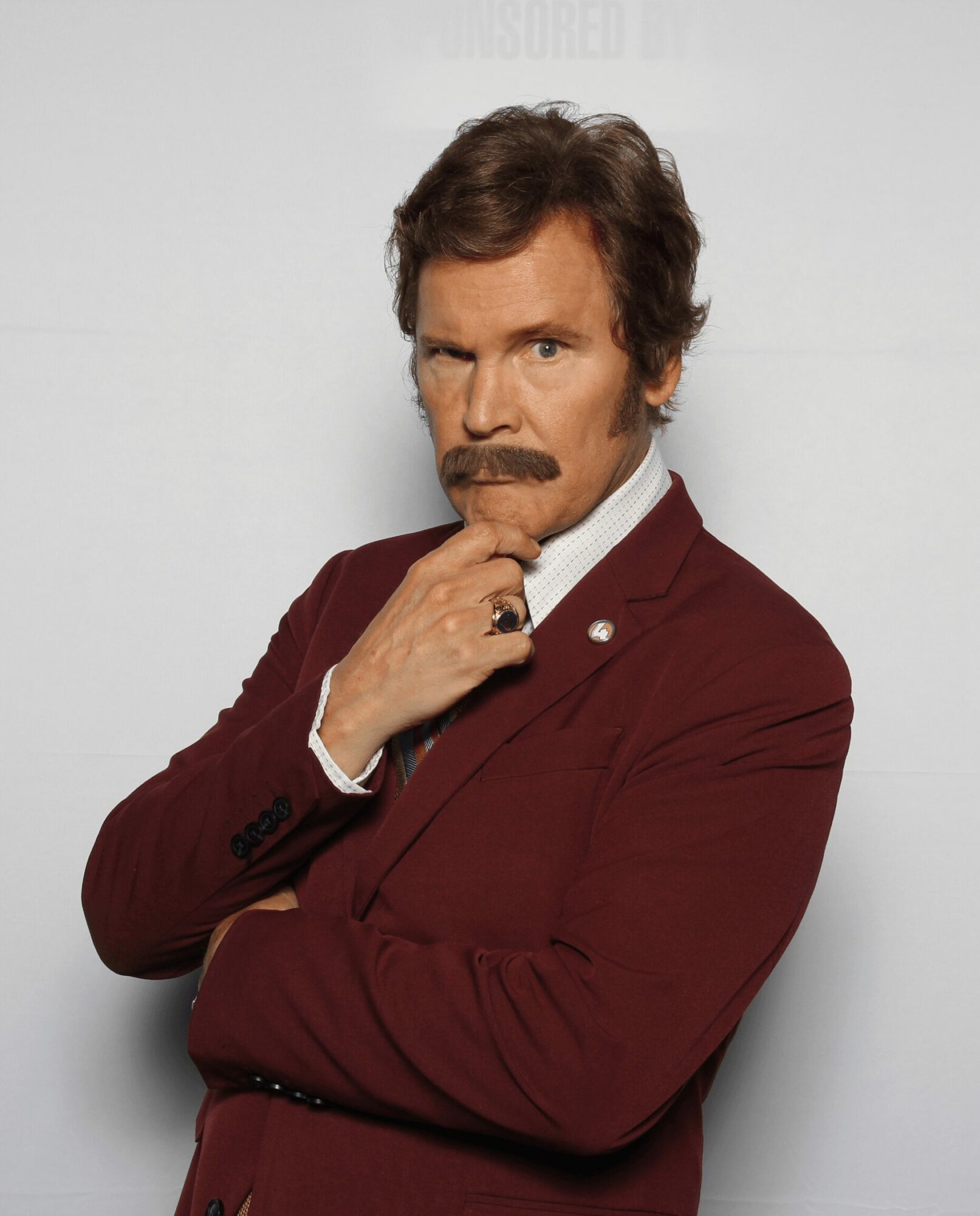 Man with brown hair and mustache in a maroon suit, posing with hand on chin, looking serious, against a plain background.