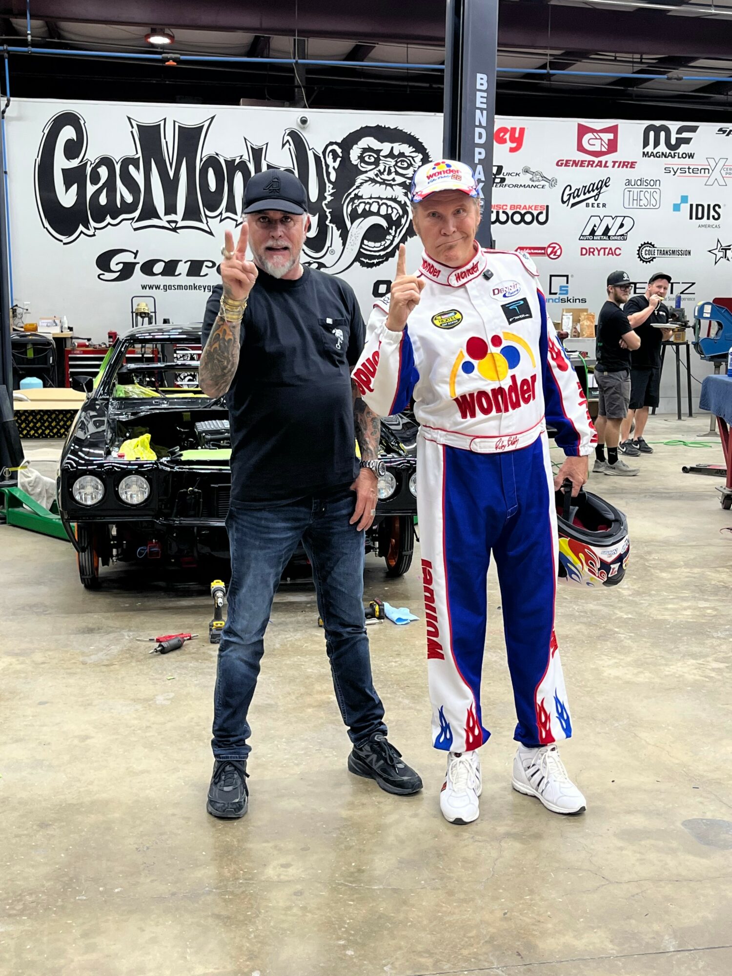 Two men standing in a racing garage area, one in casual clothing and the other in a racing suit, with race cars and banners in the background.