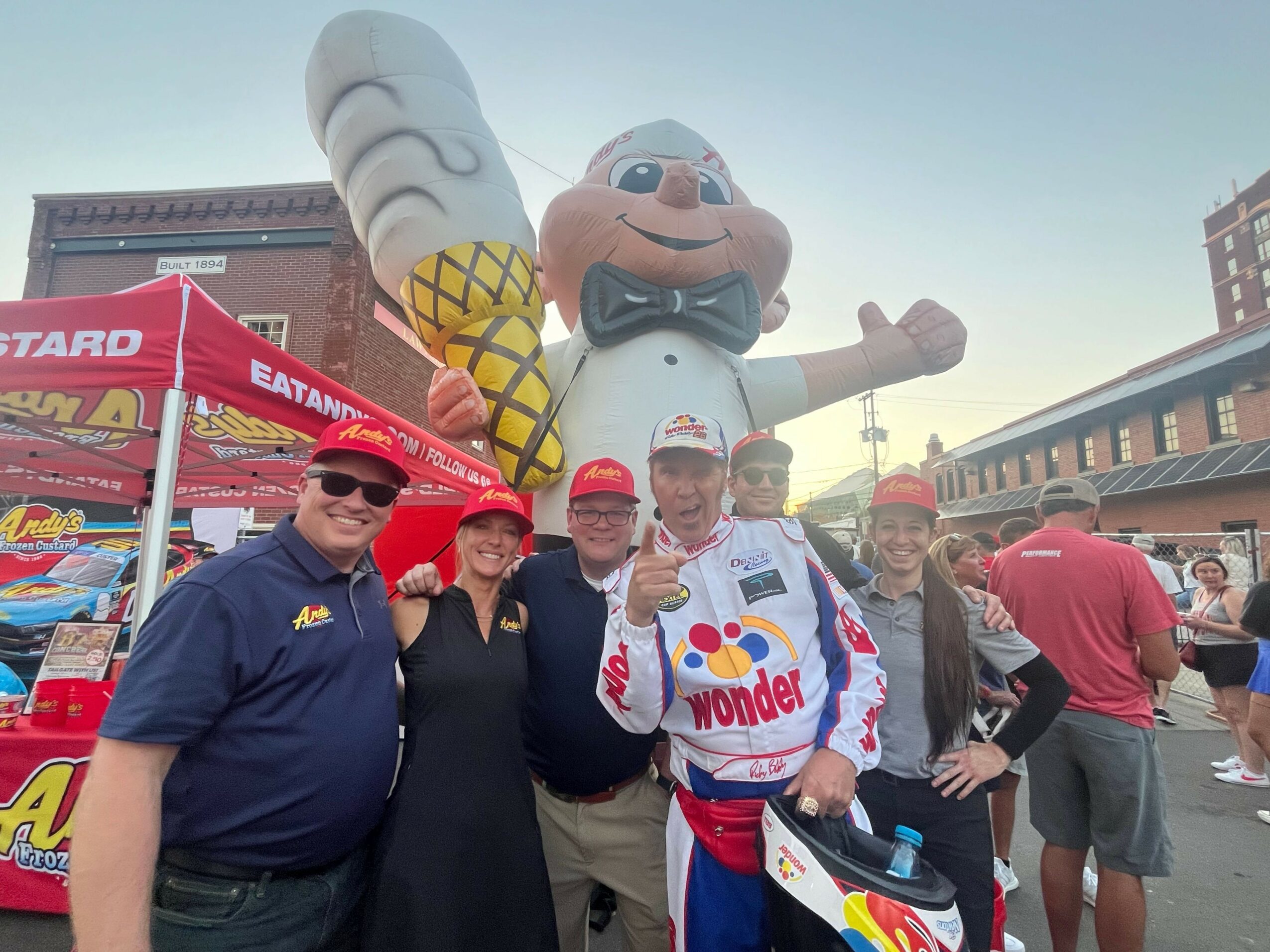 People standing outdoors near a red stand with a white and yellow mascot, some wearing sunglasses, with a clear sky.