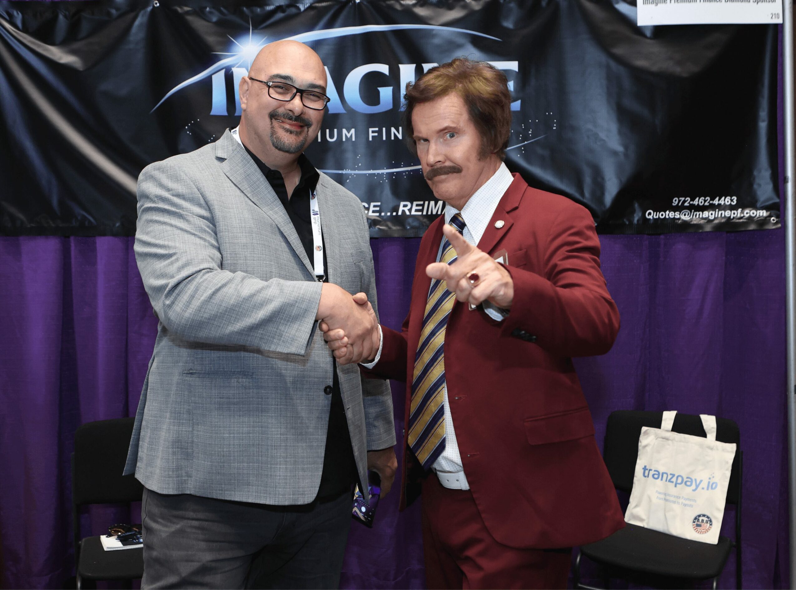 Two men shake hands, one holding a small award, in front of a purple curtain and a black banner.
