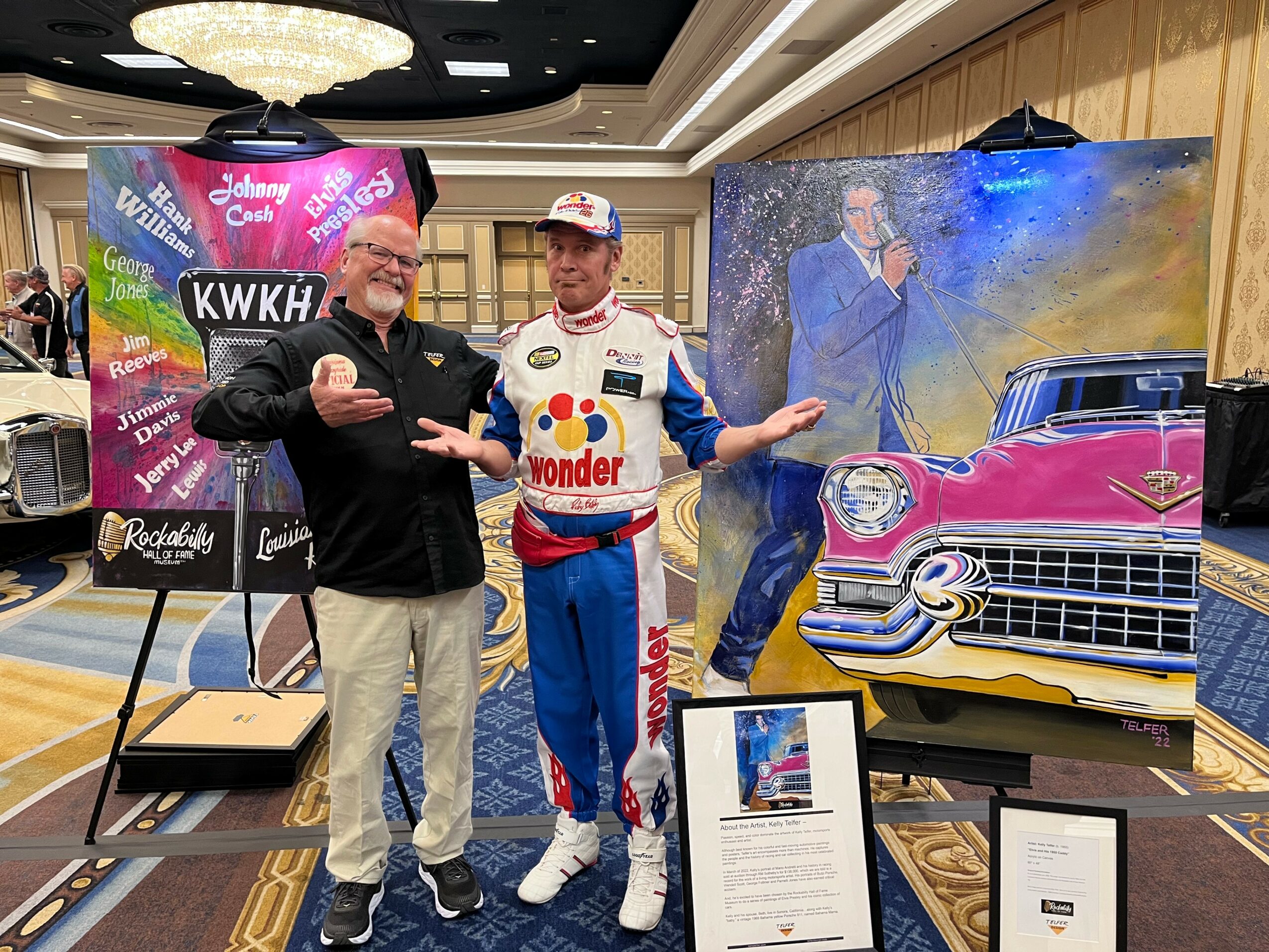 Two men stand in front of a colorful backdrop and a large painting of a pink vintage car, smiling and gesturing.