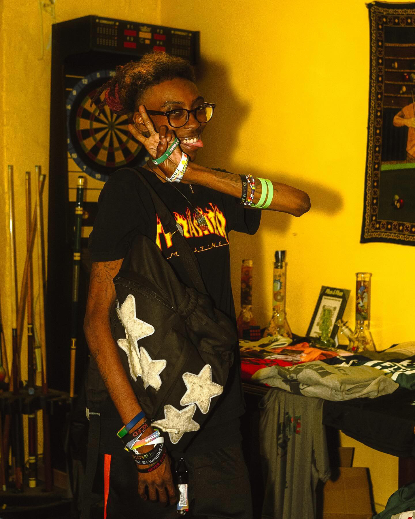 Person with glasses and curly hair smiling, wearing black clothing with butterfly designs, in a room with yellow walls and various objects.