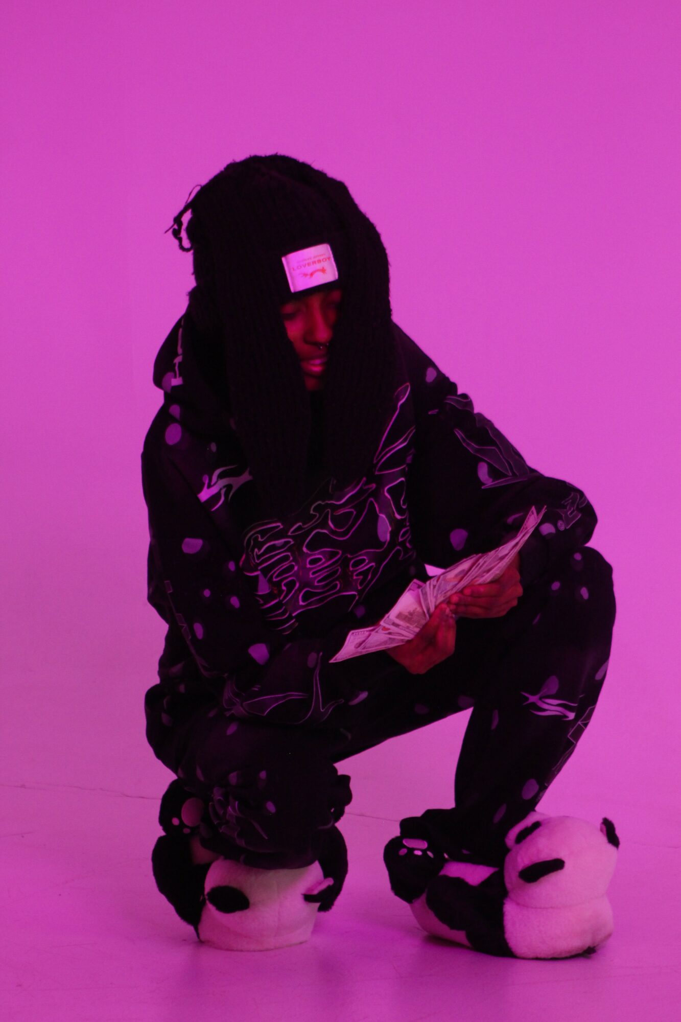 Person with dreadlocks crouching, wearing a patterned jacket and slippers, against a purple background.