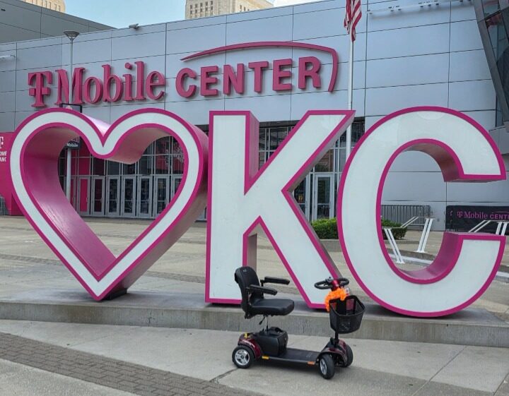 Large pink and white sign with a heart shape and the letters KC in front of a building, with a scooter nearby.