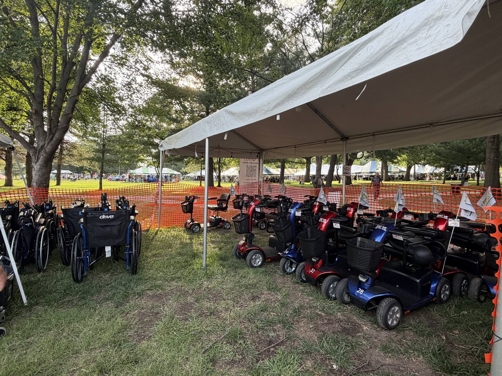 Multiple mobility scooters parked under a canopy in a grassy area with trees and orange fencing in the background.