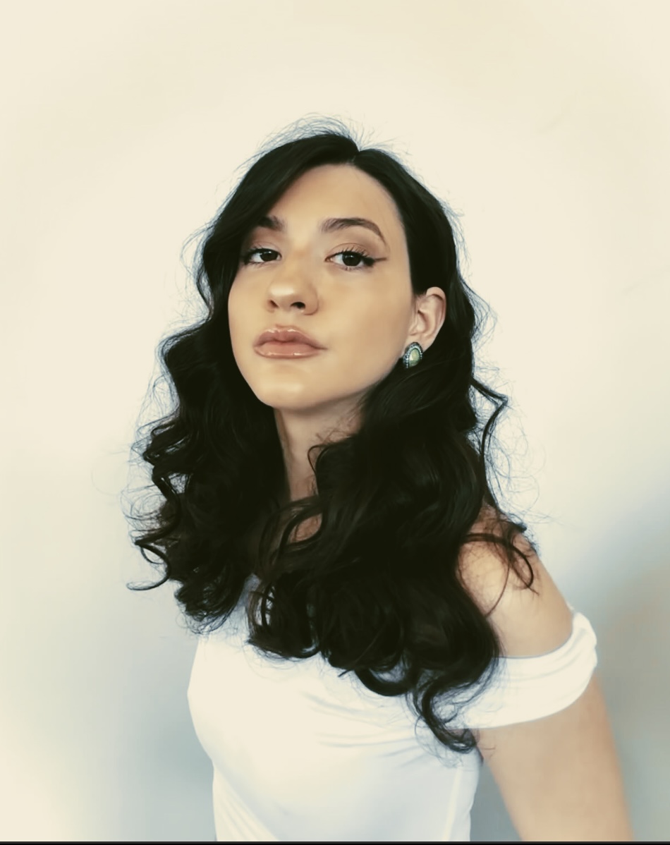 Young woman with long, wavy black hair, wearing a white off-shoulder top, looking at the camera against a plain background.