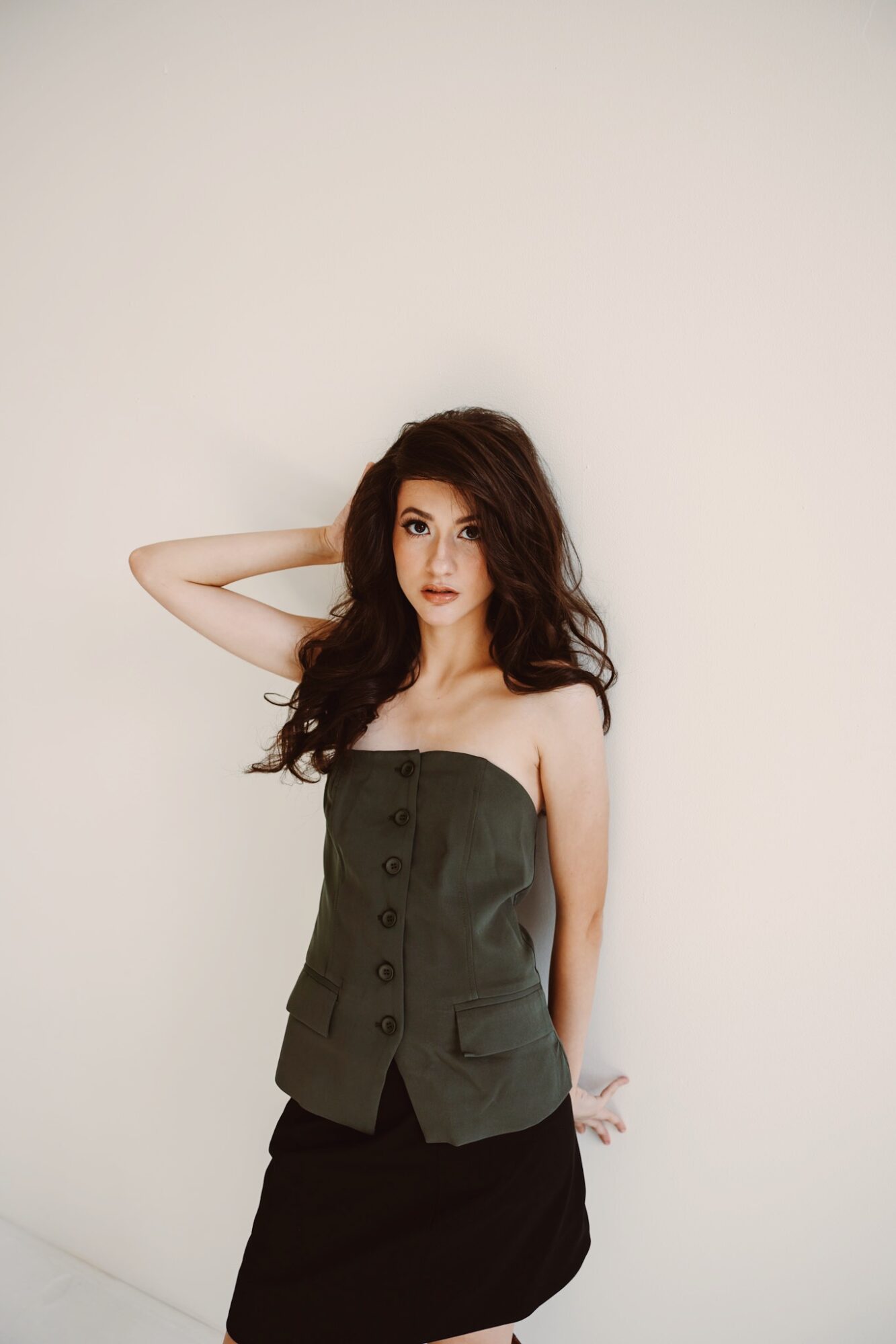 Young woman with long dark hair wearing a strapless green top and black skirt, standing against a plain white background.