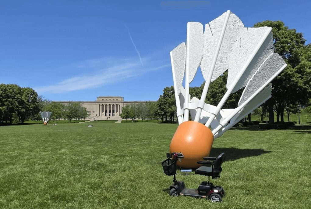 Large outdoor sculpture resembling an open book with white pages and an orange base, on a grassy field with trees and a building in the background.