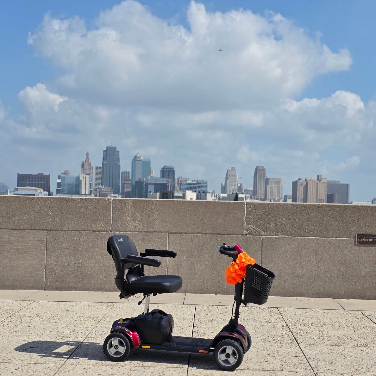City skyline with tall buildings under a partly cloudy sky, scooter with a bag and orange decoration on a sidewalk.