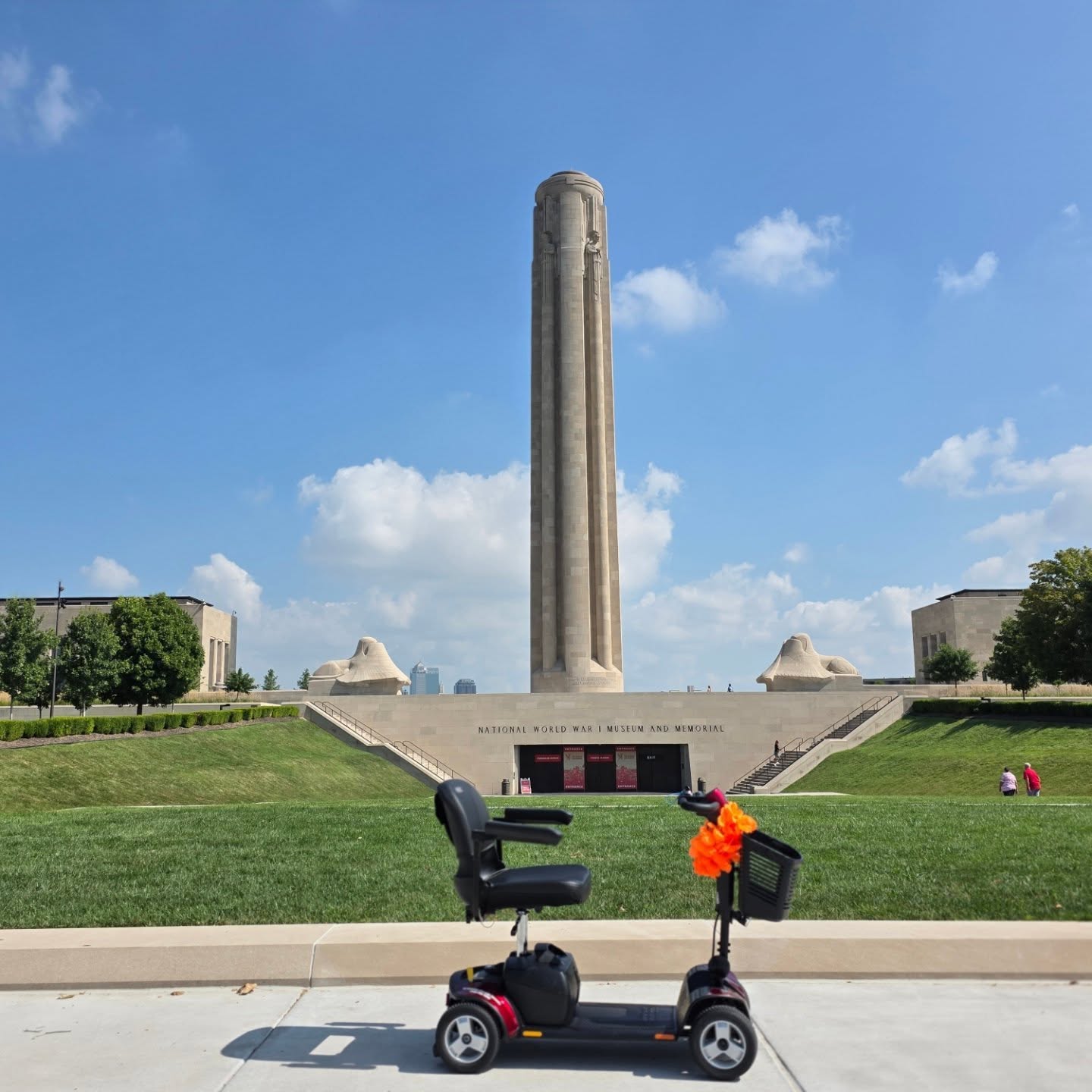 A tall monument on a grassy area with a clear blue sky, surrounded by buildings and trees.