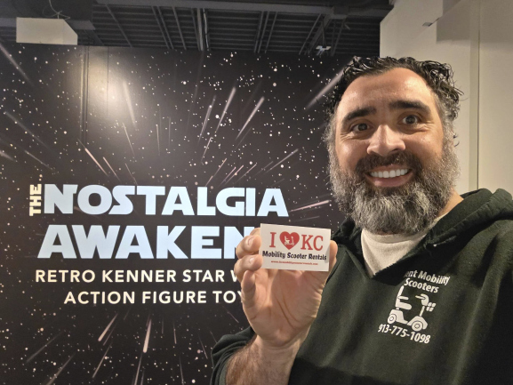 Man with a beard smiling holding a sign in front of a space-themed backdrop with text about nostalgia and retro action figures.
