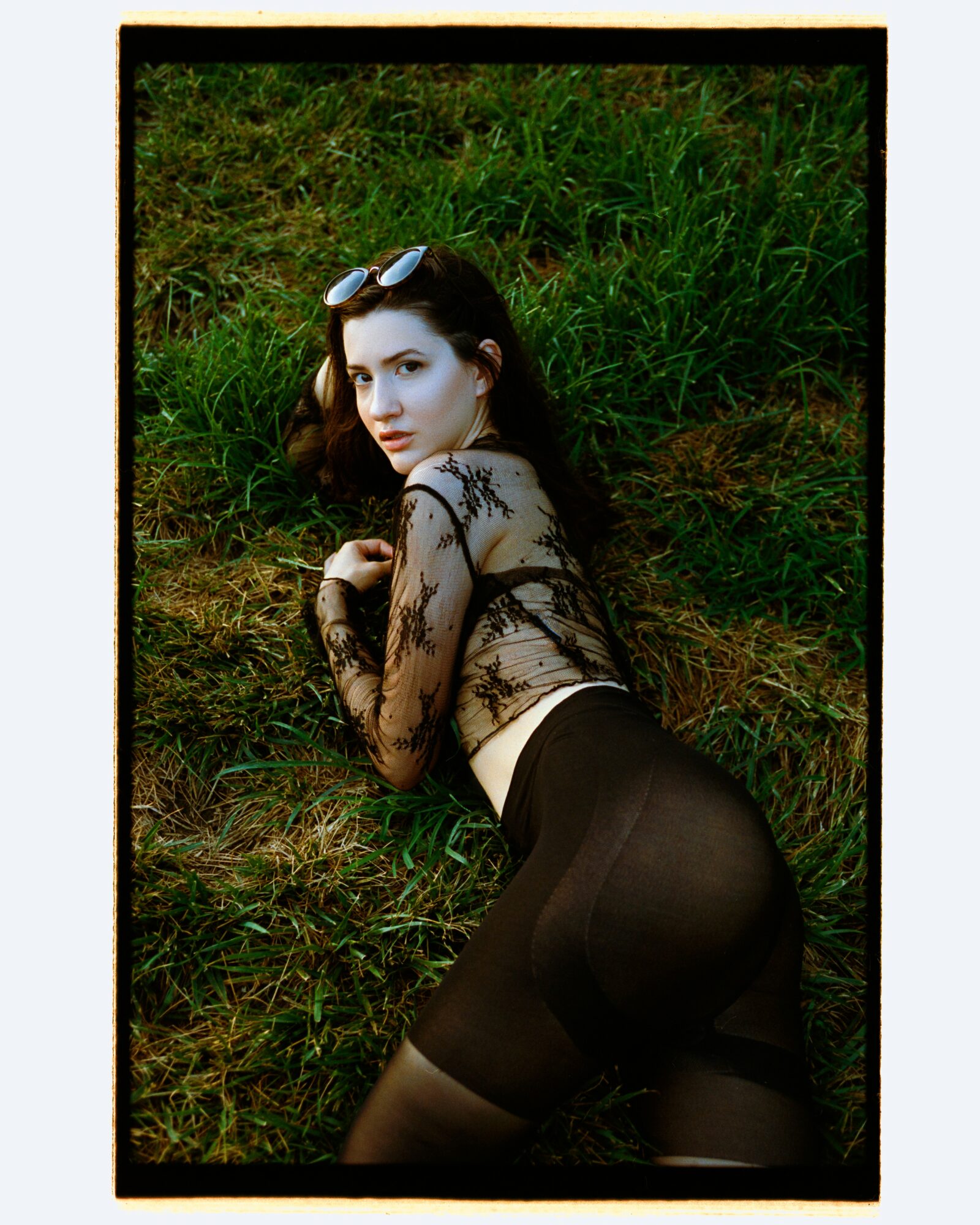 Woman lying on grass wearing a black lace top and sheer black leggings, with sunglasses on her head, looking at the camera.