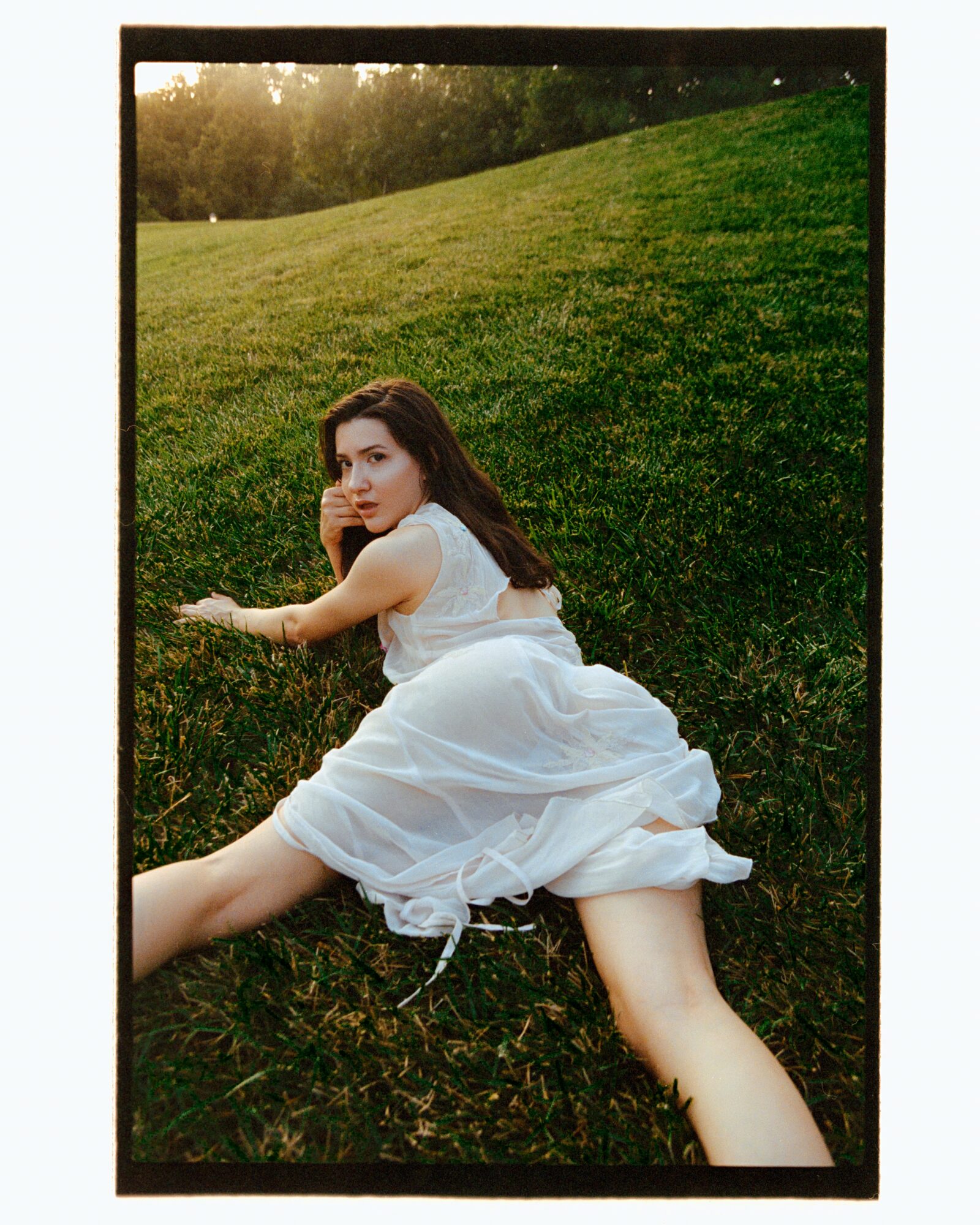 Woman in a white dress lying on grass hillside, looking at camera, with trees and sky in background.