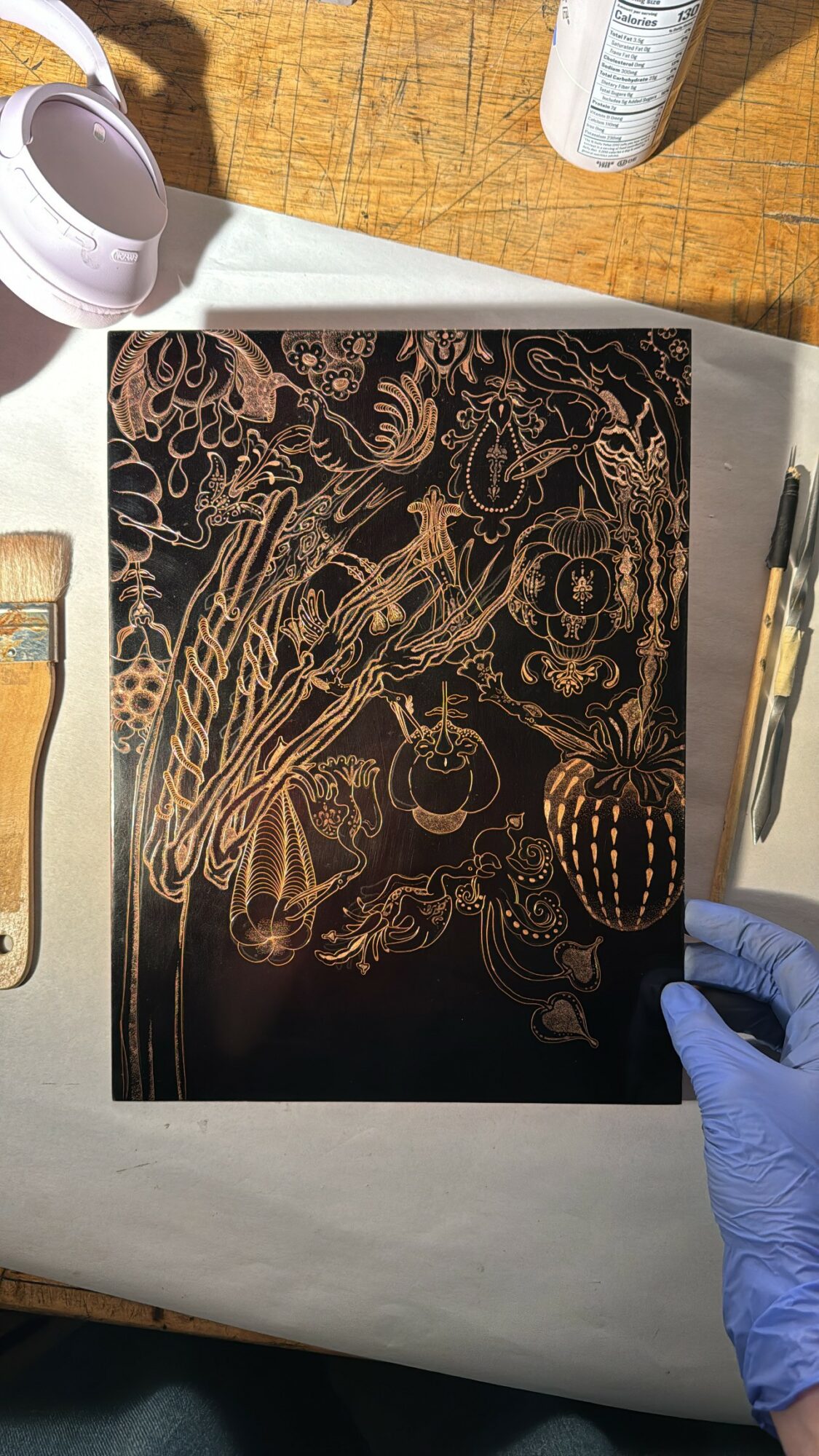 Black notebook with gold floral and abstract designs on a wooden surface, with art supplies nearby, including a paintbrush and a gloved hand.