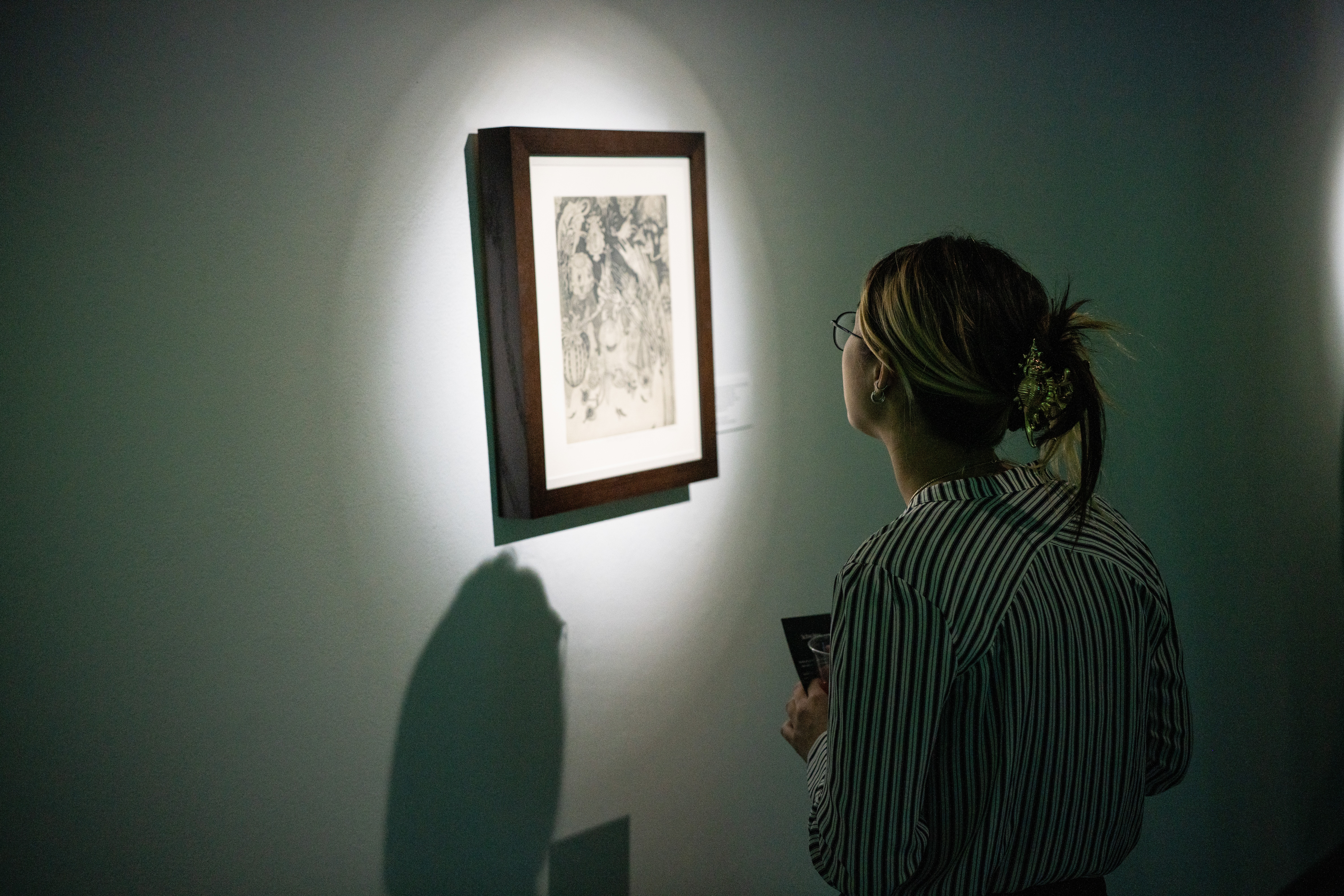Person with striped shirt and glasses viewing framed artwork on wall, holding a phone, in a dimly lit room