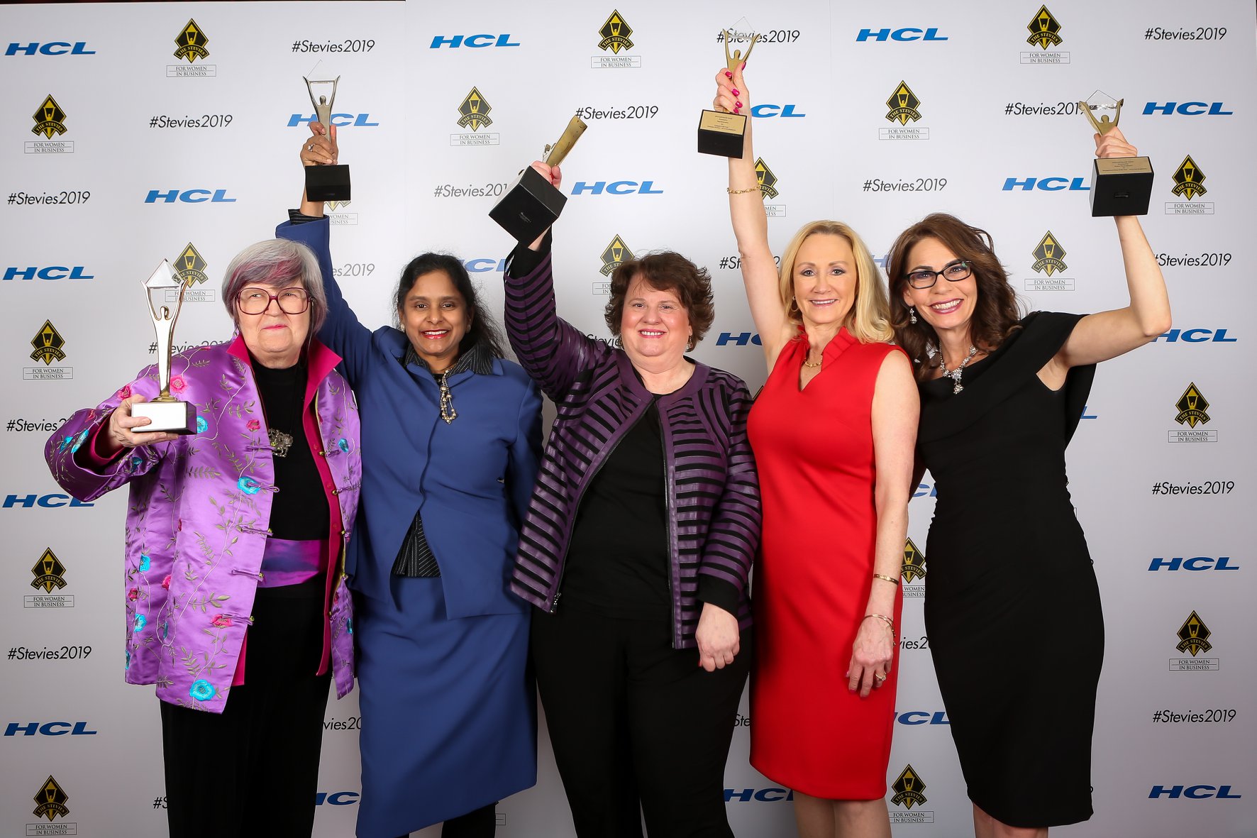 Five women standing together holding awards, smiling, in front of a backdrop with logos, celebrating achievement.