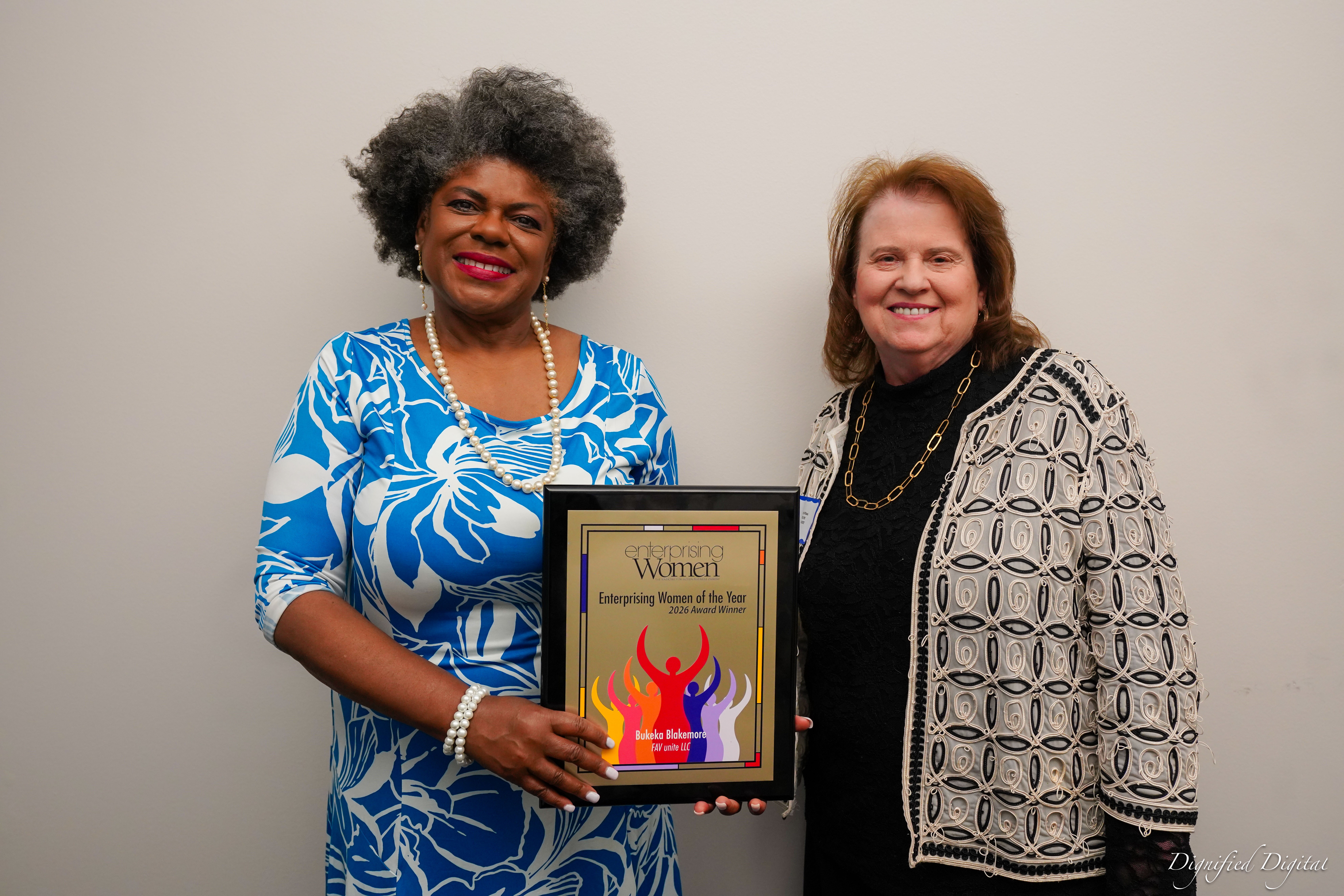 Two women smiling, holding a framed award, standing against a plain background.