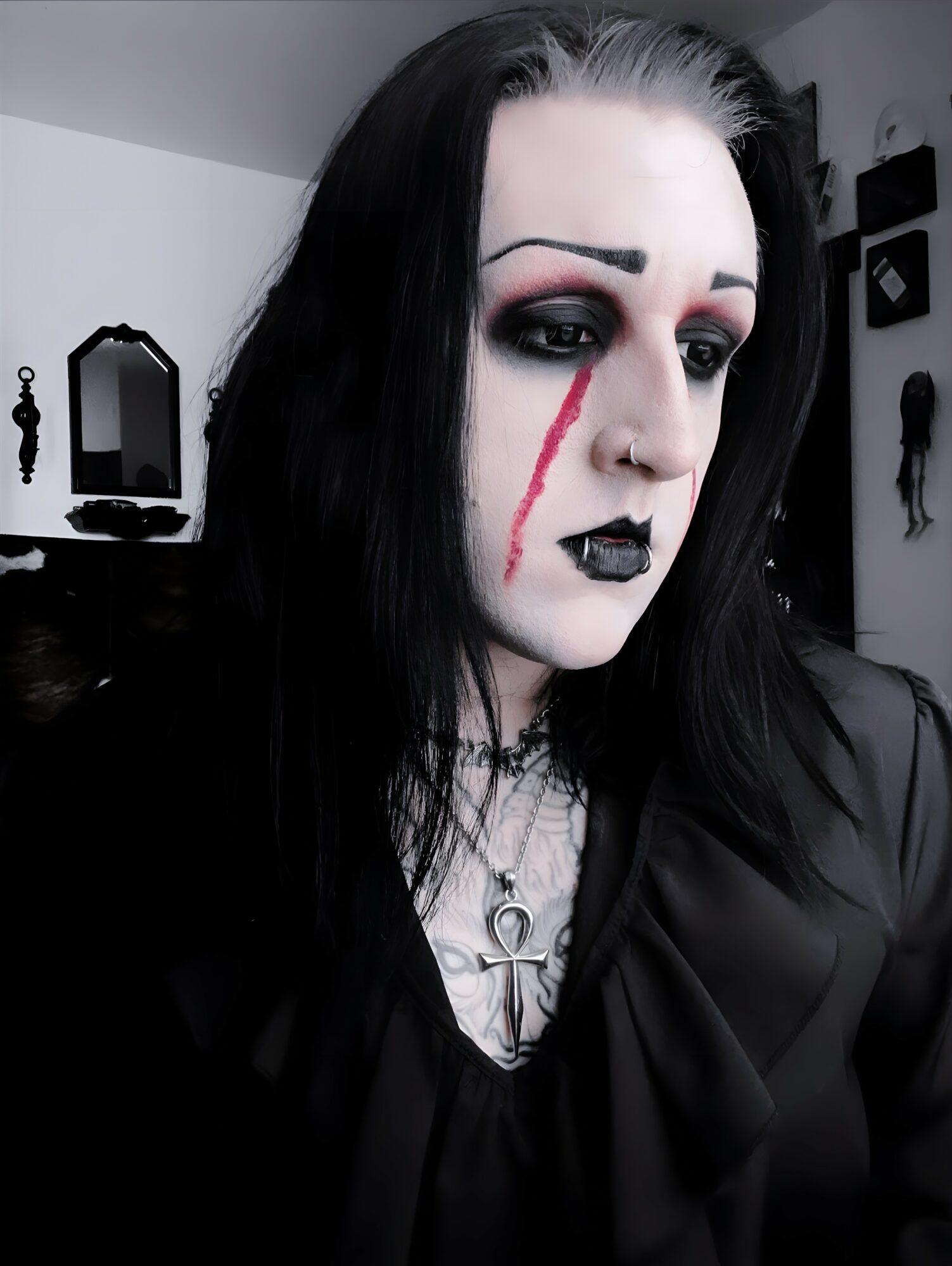 Person with black hair, dark makeup, and a red streak on face, wearing black clothing, in a room with mirrors and decorations.