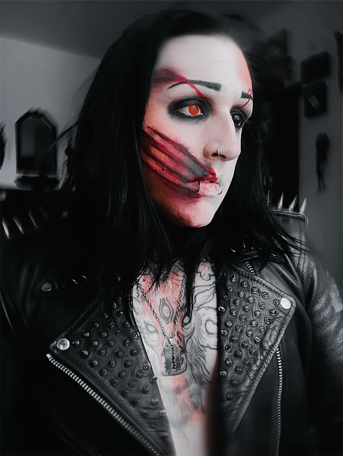 Person with face makeup resembling a stitched mouth, wearing a black leather jacket, dark hair, and tattoos visible on chest.
