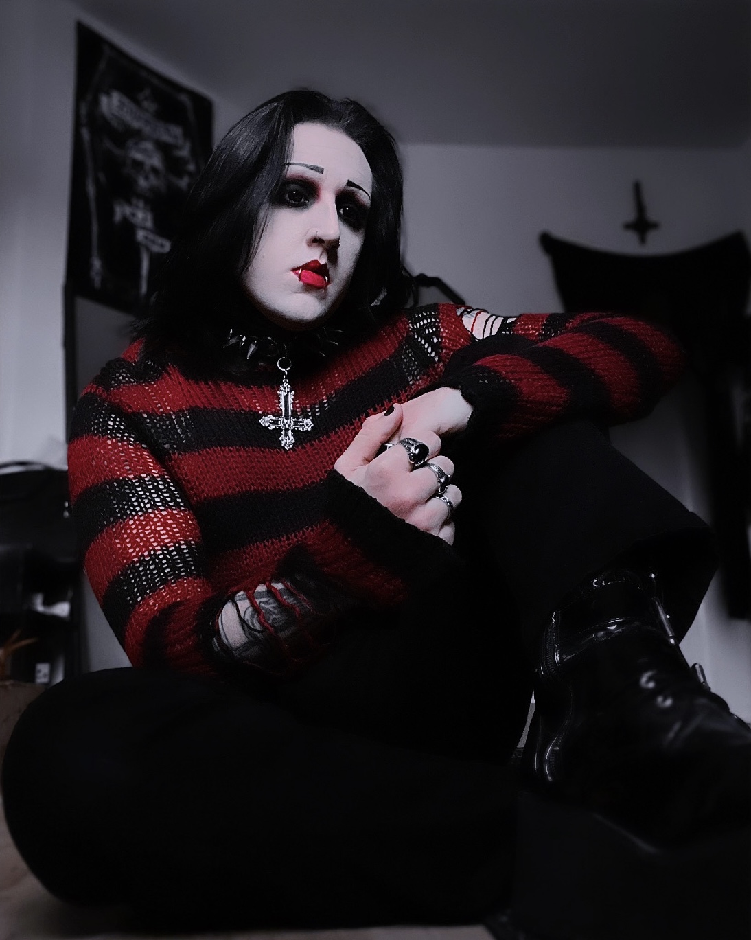 Person with dark hair, makeup, and gothic clothing sitting indoors, with a dark background and cross on the wall.