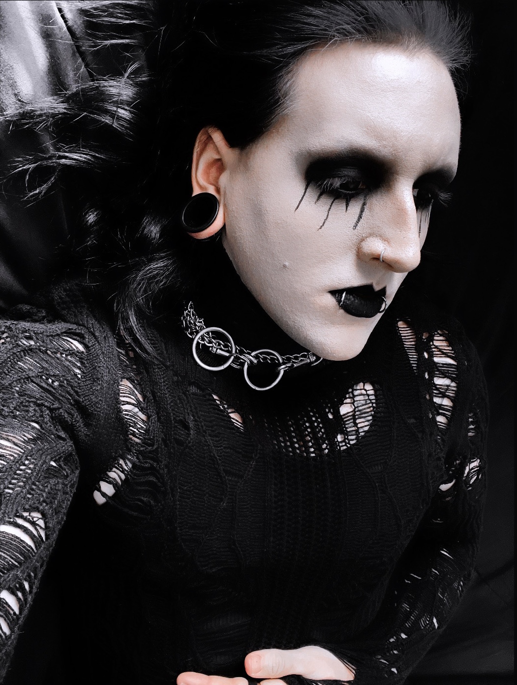 Person with dark makeup, black clothing, and gothic accessories, looking downward with a serious expression.