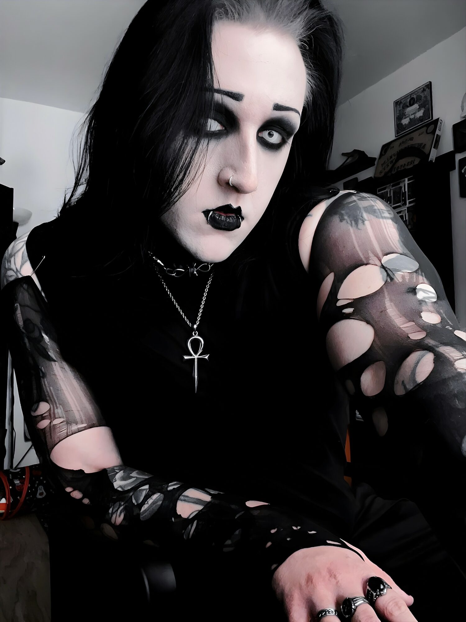Person with black hair, dark makeup, and gothic clothing, including ripped sleeves and an ankh necklace, in a room.