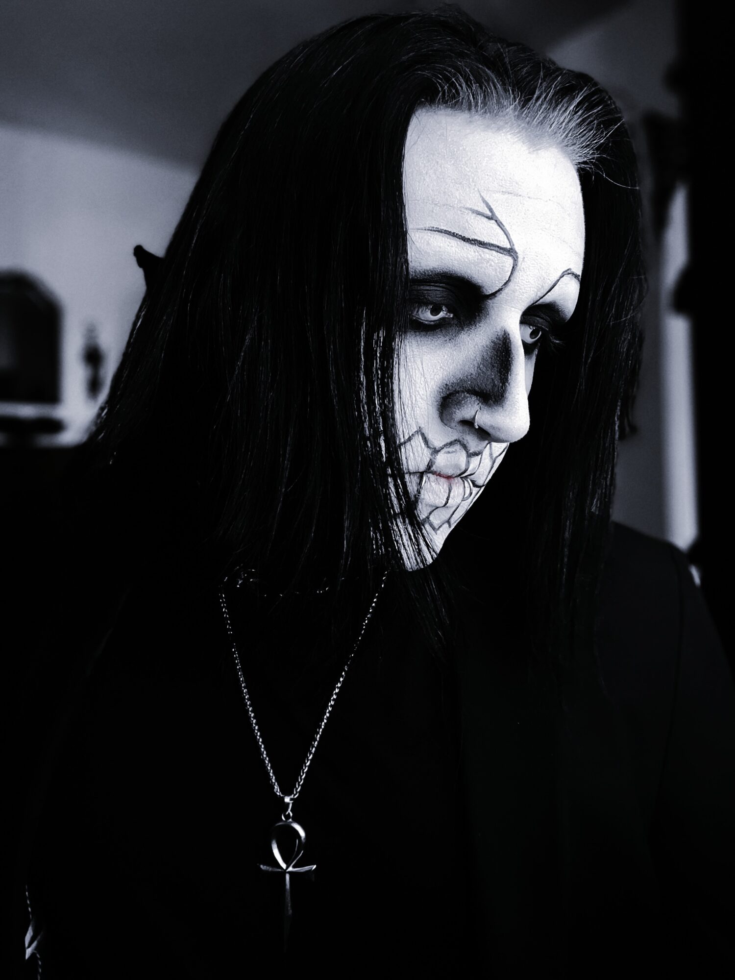 Person with skull makeup, dark hair, and an ankh necklace, looking to the side in black and white.
